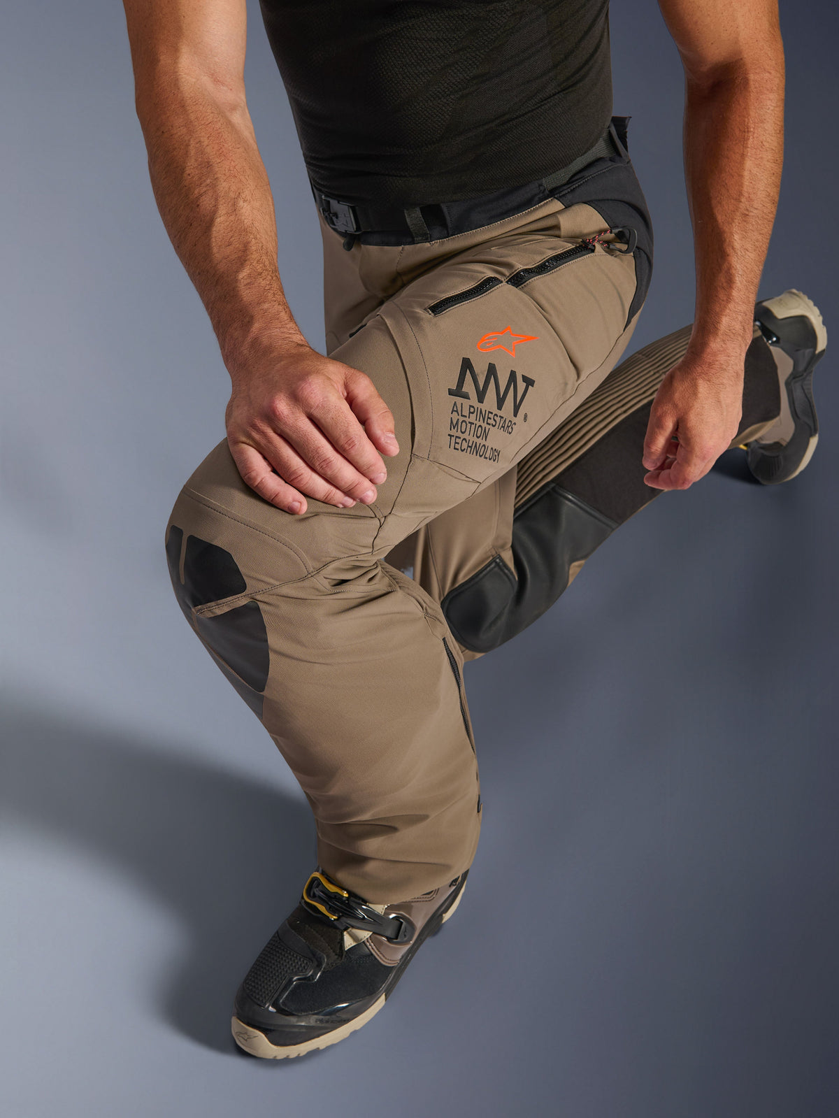 Pantalon Alpinestars AMT-8 Stretch Drystar® XF en Walnut Black, avec le marquage Alpinestars Motion Technology (AMT) sur la cuisse, de longs ports de ventilation zippés et des panneaux de genoux en cuir durables, montré sur un pilote en position agenouillée pour souligner la flexibilité.