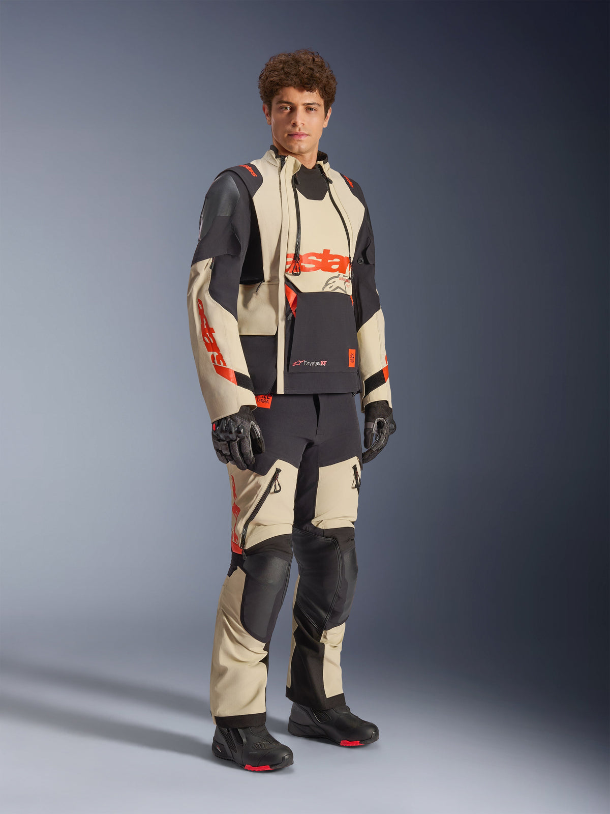 Modèle masculin portant la veste laminée Alpinestars Halo Pro Drystar® XF en Mastic Black, présentant des panneaux beiges et noirs avec des accents rouges, associée à un pantalon d'aventure assorti et des gants noirs, conçue pour la conduite de moto d'aventure tout-terrain.