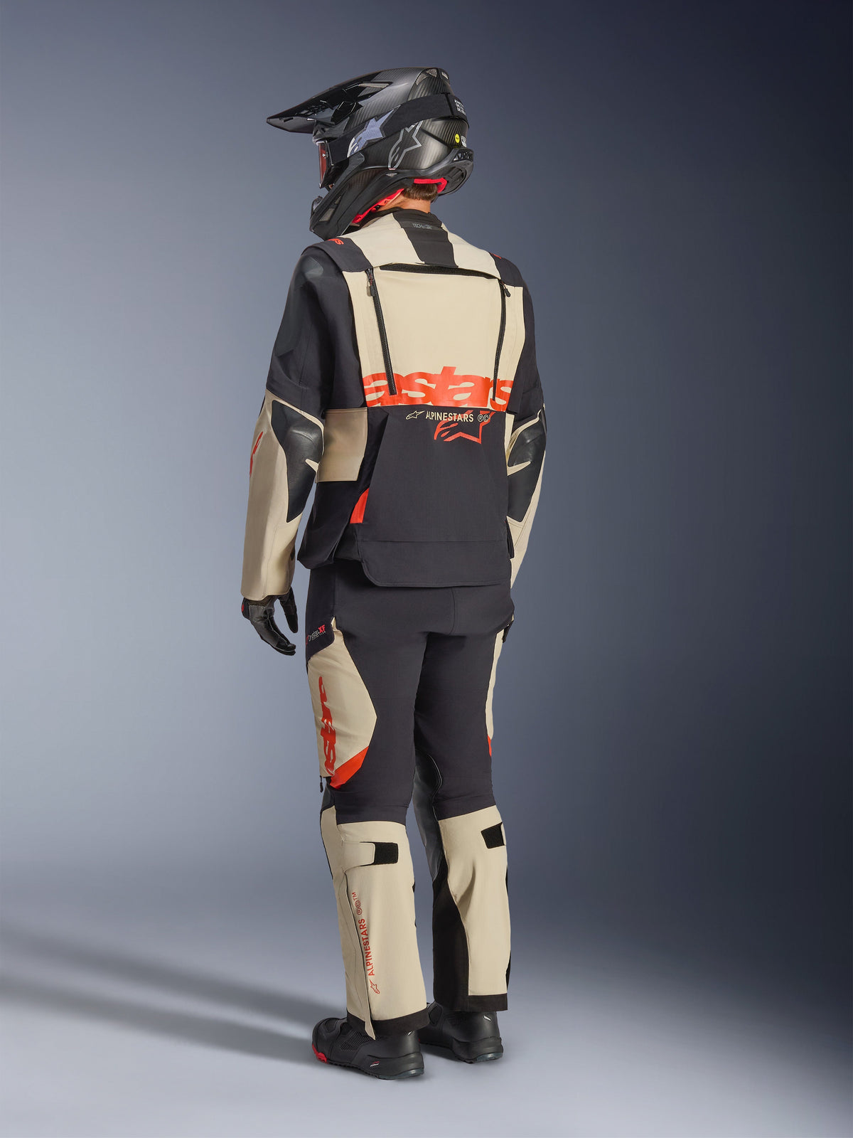 Veste laminée Alpinestars Halo Pro Drystar® XF, Mastic Black, vue arrière d'un pilote portant la tenue de moto d'aventure complète avec pantalon assorti, casque d'aventure noir et bottes, arborant un logo Alpinestars rouge vif imposant dans le dos.