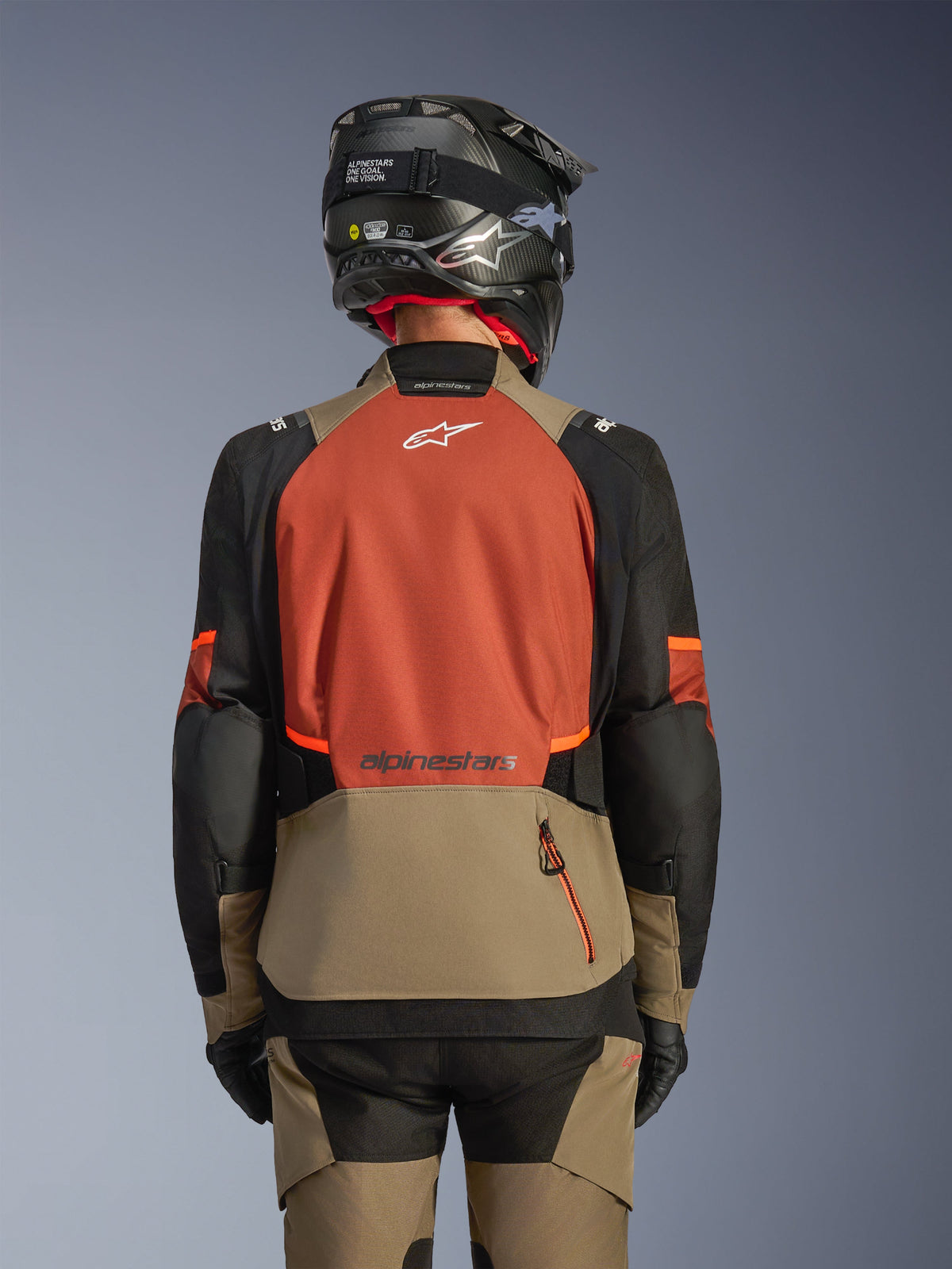 Vue arrière de la veste Alpinestars Andes V4 Drystar® en Walnut Rust Black, veste d'aventure touring dotée d'un grand panneau arrière de couleur rouille avec un logo blanc, d'une section inférieure noyer avec une poche zippée verticale, de manches noires avec des accents orange, portée avec un pantalon assorti et un casque noir pour une conduite par tous les temps.