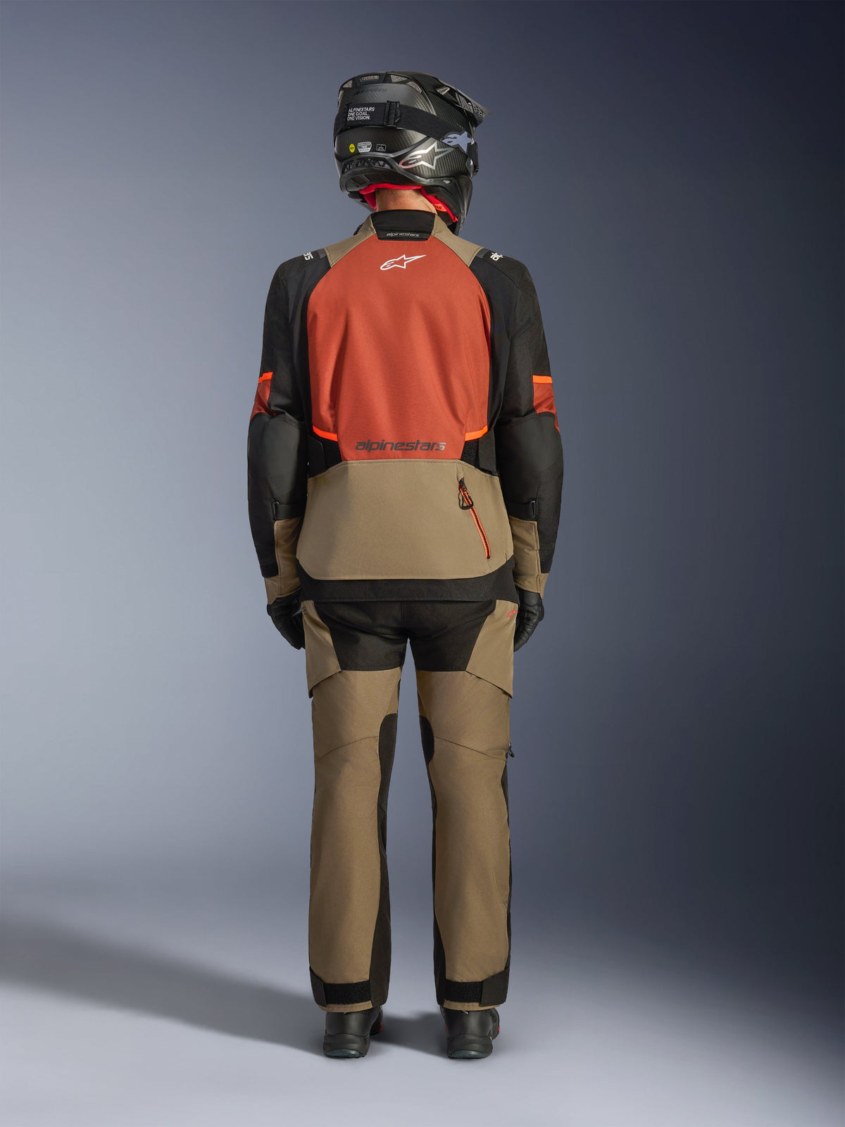 Vue arrière d'un pilote portant la veste Alpinestars Andes V4 Drystar® en Walnut Rust Black, associée à un pantalon d'aventure walnut assorti et un casque noir, mettant en valeur le panneau arrière de couleur rouille, la grande poche de rangement arrière et les logos réfléchissants de la veste.