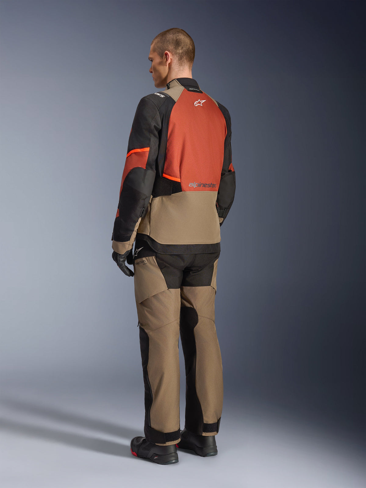 Veste Alpinestars Andes V4 Drystar® en Walnut Rust Black, modèle vu de dos portant un pantalon en textile assorti, des gants noirs et des bottes, présentant une conception technique multi-panneaux pour le touring d'aventure et le logo Alpinestars.