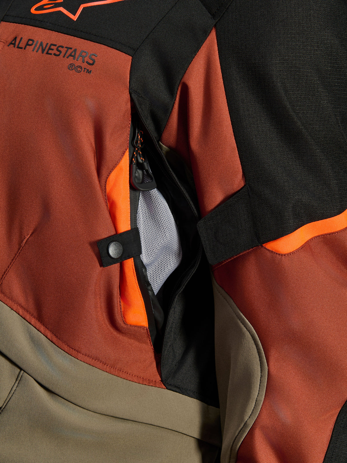 Veste Alpinestars Andes V4 Drystar®, Walnut Rust Black, gros plan sur l'ouverture de ventilation directe avec doublure en maille, textile durable avec accents orange, bouton-pression et détail du logo Alpinestars
