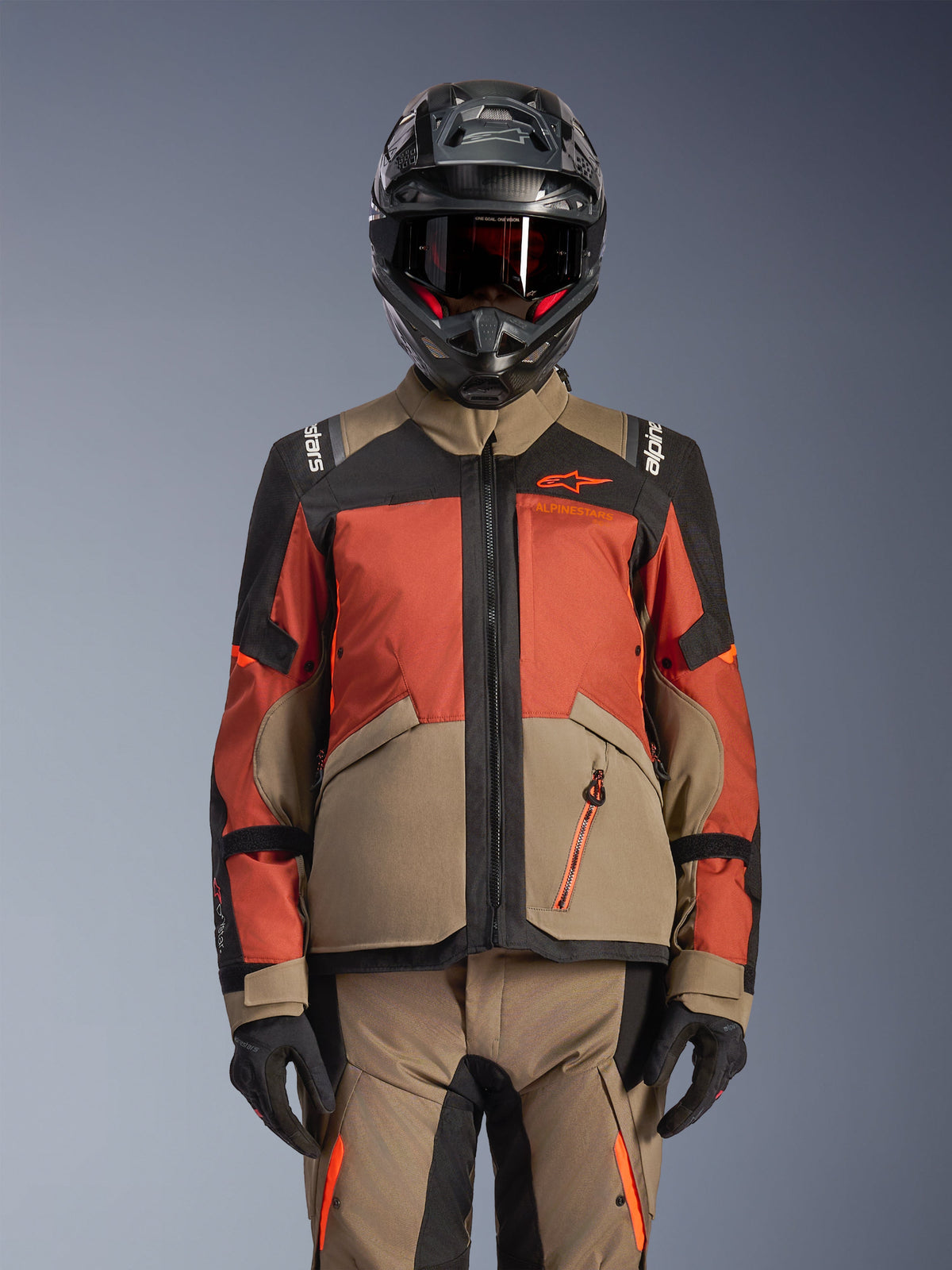 Veste Alpinestars Andes V4 Drystar®, veste de moto d'aventure, Walnut Rust Black, portée avec un casque et des gants assortis, dotée d'une conception technique multi-panneaux en beige, rouge orangé et noir avec logos orange visibles et fermetures éclair imperméables.