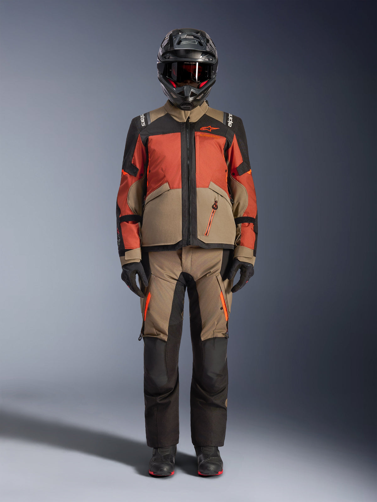 Motard d'aventure portant la veste Alpinestars Andes V4 Drystar® en Walnut Rust Black, associée à un pantalon d'aventure assorti, un casque noir avec des accents rouges et des lunettes, et des gants noirs, présentant une tenue de touring complète toutes saisons.