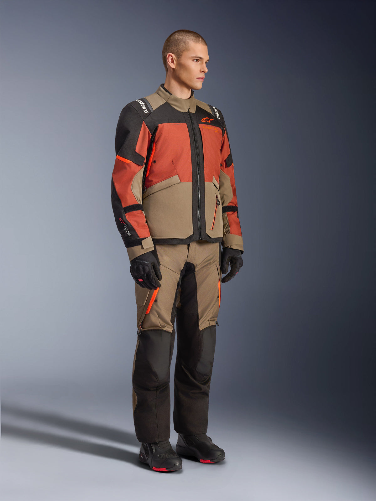 Veste Alpinestars Andes V4 Drystar®, veste d'aventure touring, noyer, rouille et noir, portée avec un pantalon assorti et des bottes noires, présentant un design à blocs de couleurs en beige, rouille et noir, construction textile durable avec fermetures éclair visibles et logos Alpinestars.