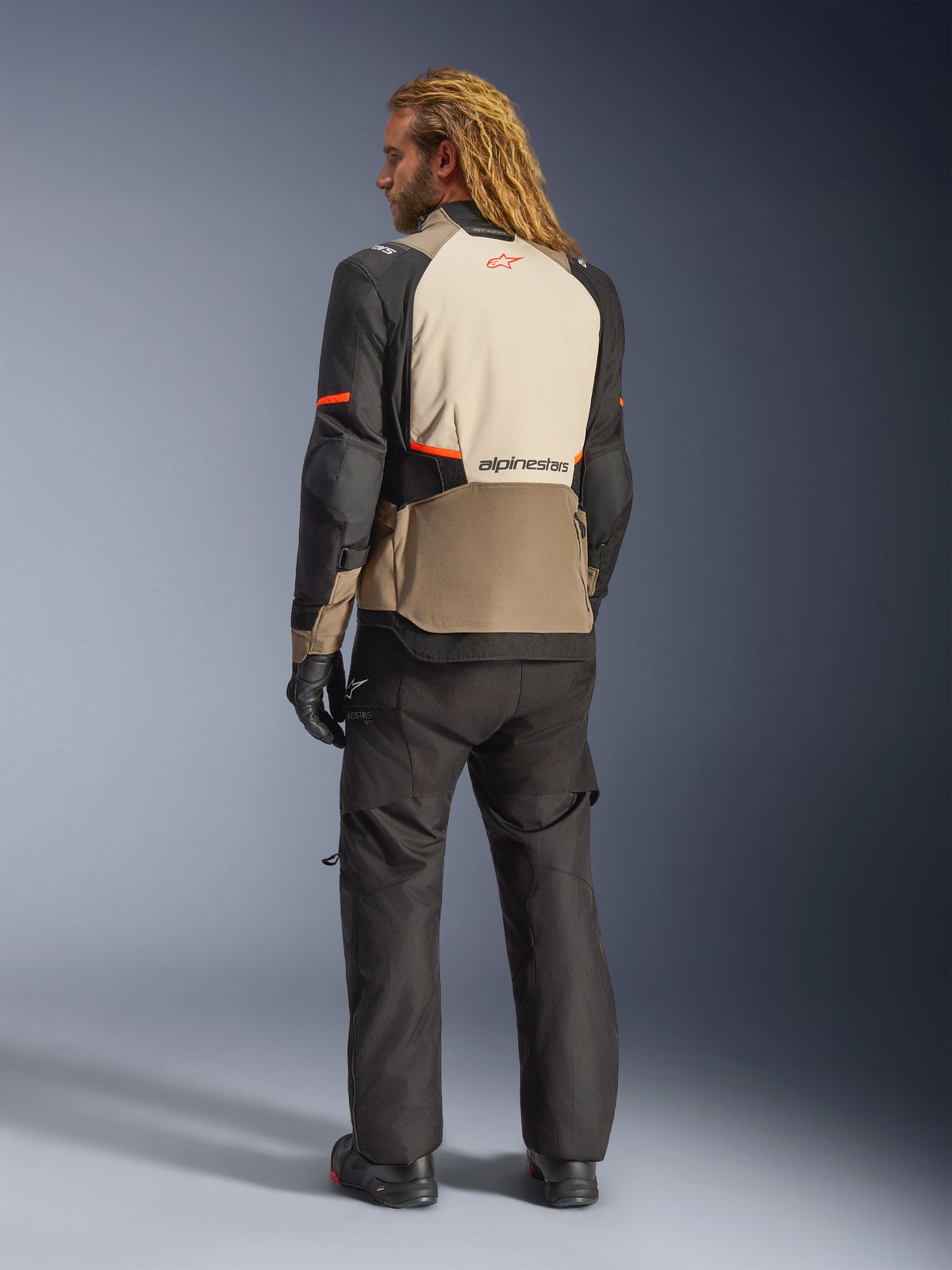 Veste Alpinestars Andes V4 Drystar®, Walnut Mastic Black, vue arrière sur un modèle masculin, présentant des panneaux textiles renforcés beige et noir avec des touches d'orange, logo Alpinestars dans le dos, portée avec un pantalon de moto d'aventure noir et des gants assortis.