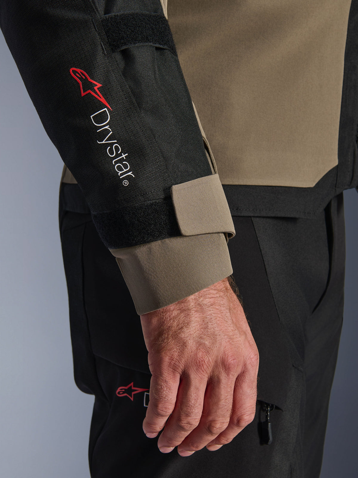 Gros plan sur la veste Alpinestars Andes V4 Drystar® en coloris Walnut et Noir, avec le logo Drystar® rouge sur l'avant-bras et une sangle de poignet réglable, faisant partie d'une tenue de moto d'aventure technique.