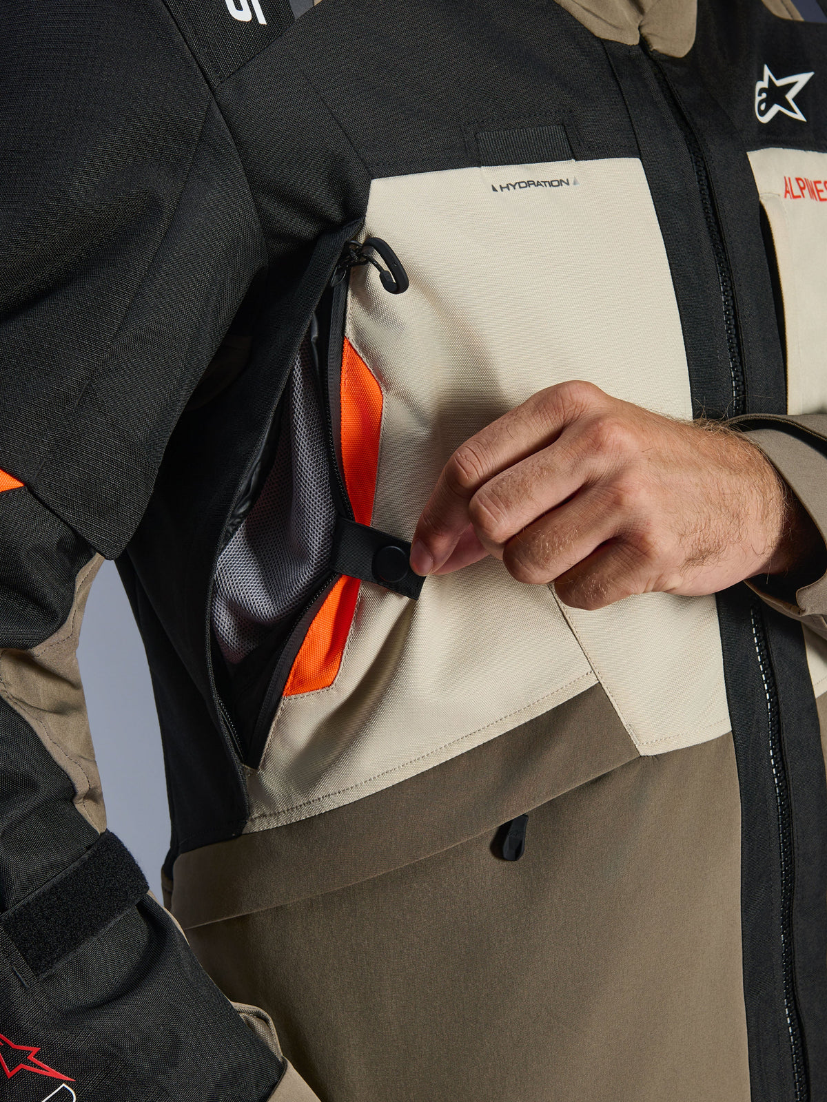 Veste Alpinestars Andes V4 Drystar®, vue détaillée, textile Noyer Mastic Noir, gros plan d'une main ouvrant le panneau de ventilation sur la poitrine pour révéler un intérieur en mesh orange vif, montrant le système de flux d'air direct pour la conduite d'aventure