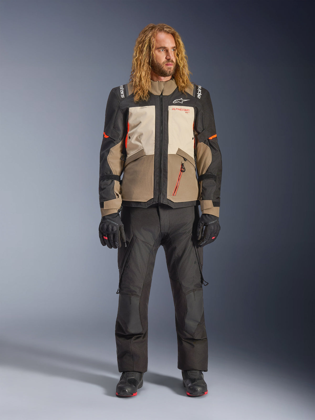Veste Alpinestars Andes V4 Drystar®, Walnut Mastic Black, portée avec un pantalon de moto et des gants noirs, design aventure multi-panneaux avec accents orange et logo Alpinestars, équipement textile technique pour rouler par tous les temps