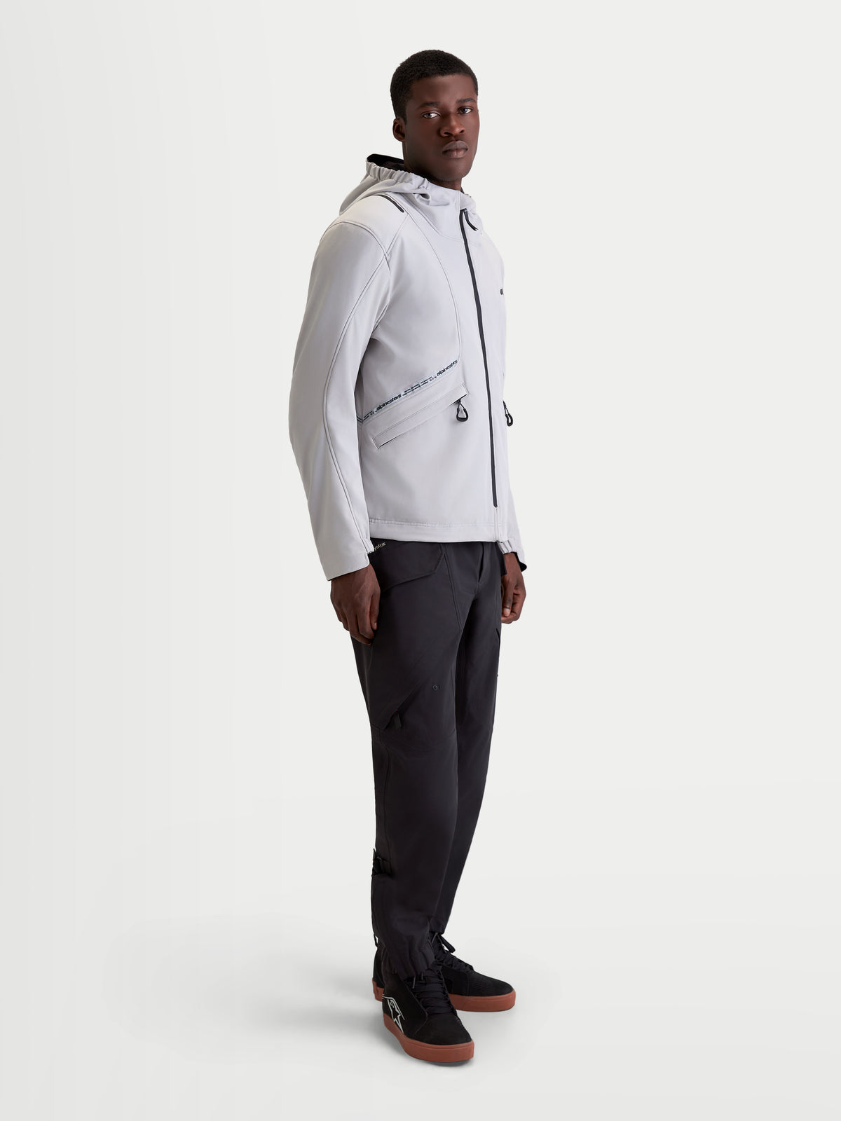 Alpinestars C 5 Drystar Veste, veste de moto urbaine contemporaine pour homme en gris clair et noir, vue de profil sur un modèle portant un pantalon sombre assorti et des chaussures de moto, construction softshell 2 couches imperméable avec capuche intégrée