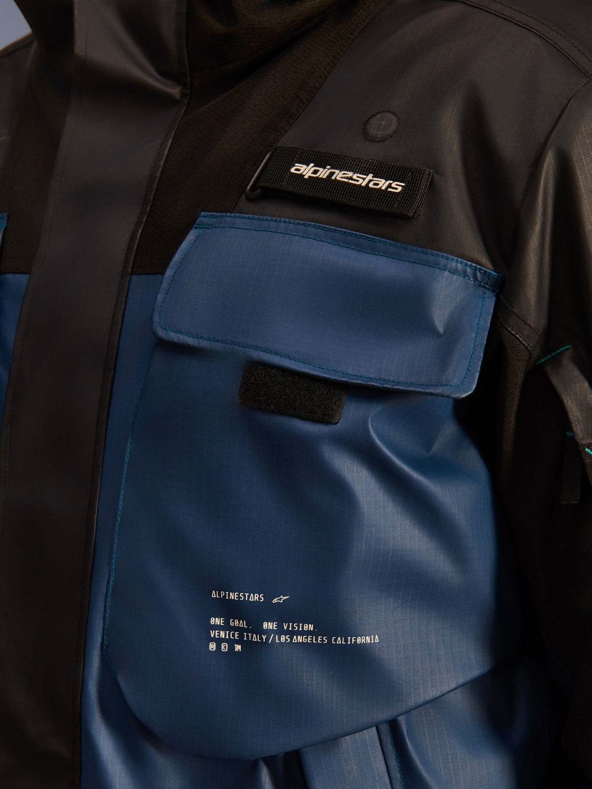 Veste imperméable Alpinestars Mospher, gros plan sur la poche de poitrine, Noir et Dark Petrol, tissu ripstop résistant avec marquage imprimé « One Goal. One Vision. », design utilitaire avec poches fonctionnelles et languette logo Alpinestars