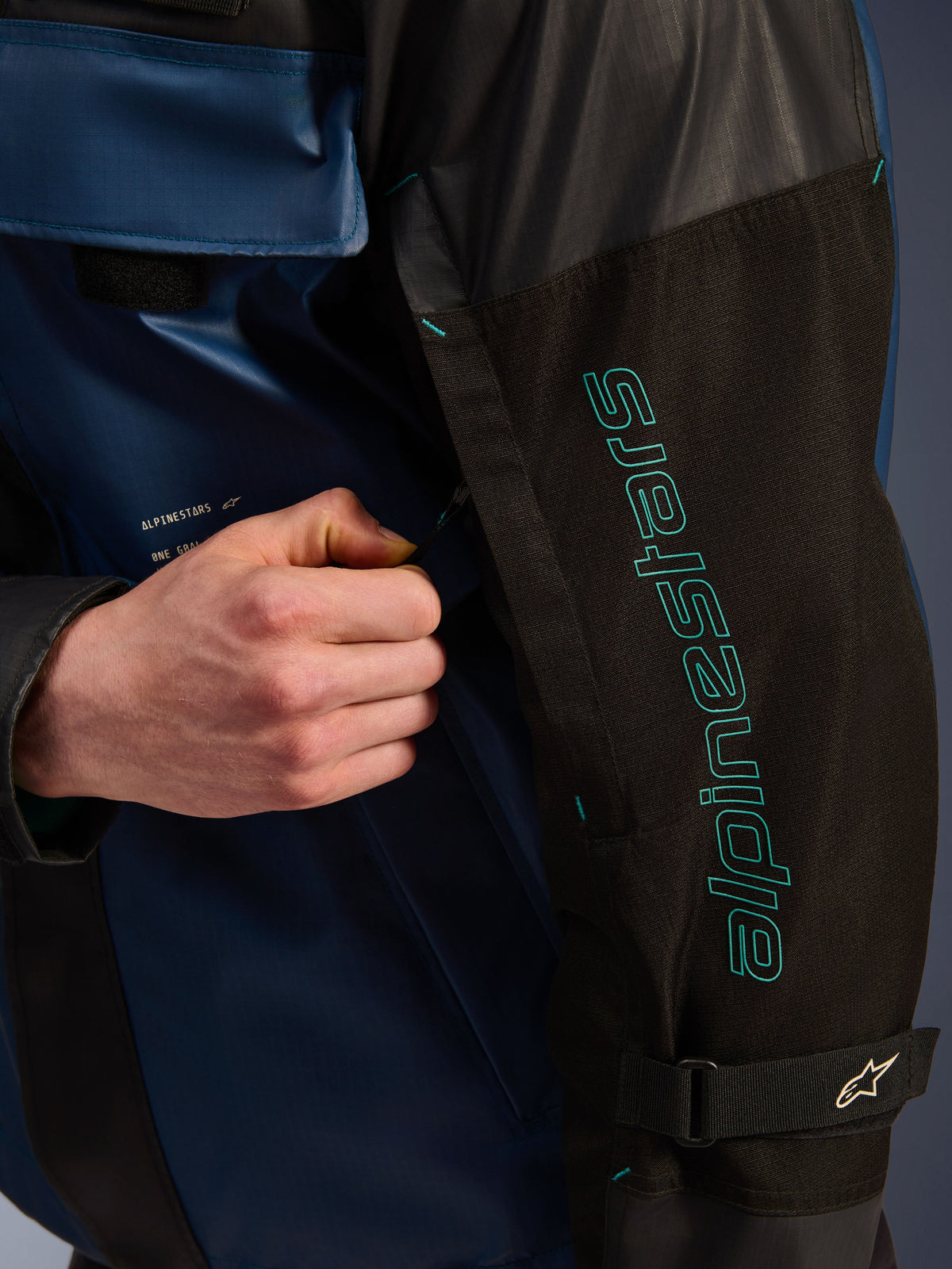 Veste imperméable Alpinestars Mospher en Black Dark Petrol, gros plan sur le détail de la manche avec marquage Alpinestars bleu sarcelle, une main ajustant le zip de ventilation, dotée d'un tissu ripstop durable et d'une sangle de bras réglable