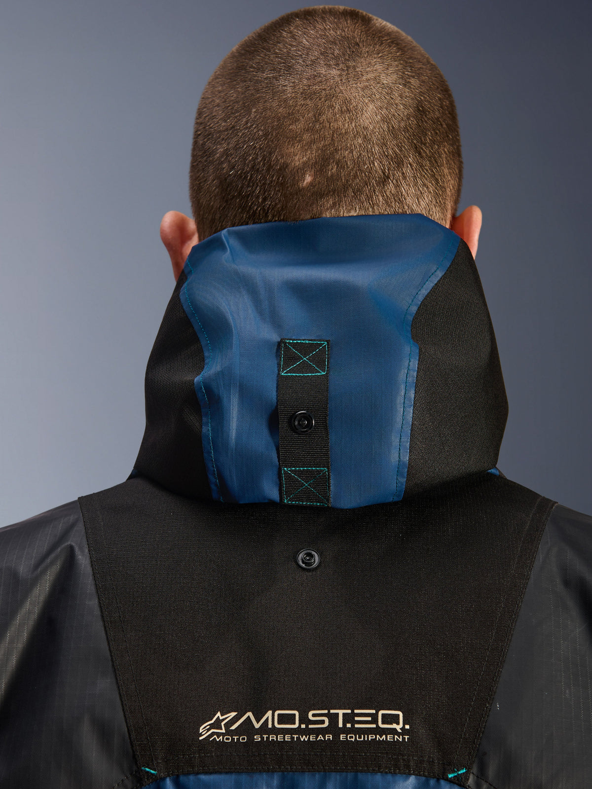 Veste Alpinestars Mospher Waterproof, vue arrière détaillée montrant le système d'ancrage de la capuche, Noir et Bleu Pétrole Foncé, avec marquage MO.ST.EQ. Moto Streetwear Equipment, textile ripstop durable, équipement moto technique urbain