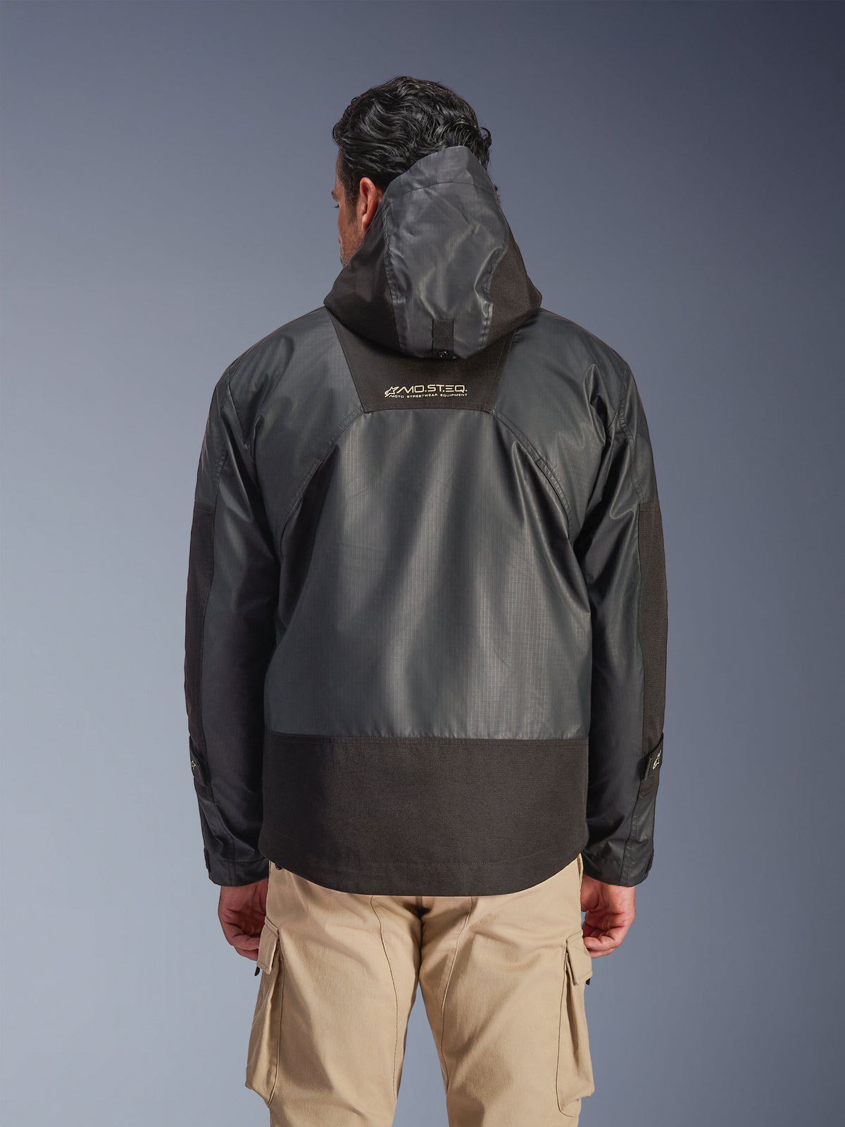Vue arrière d'un homme portant la veste Alpinestars Mospher Waterproof noire, dotée d'une capuche et d'une construction en textile ripstop durable, associée à un pantalon cargo beige, mettant en avant le logo MO.ST.EQ. dans le haut du dos, design d'équipement moto urbain.