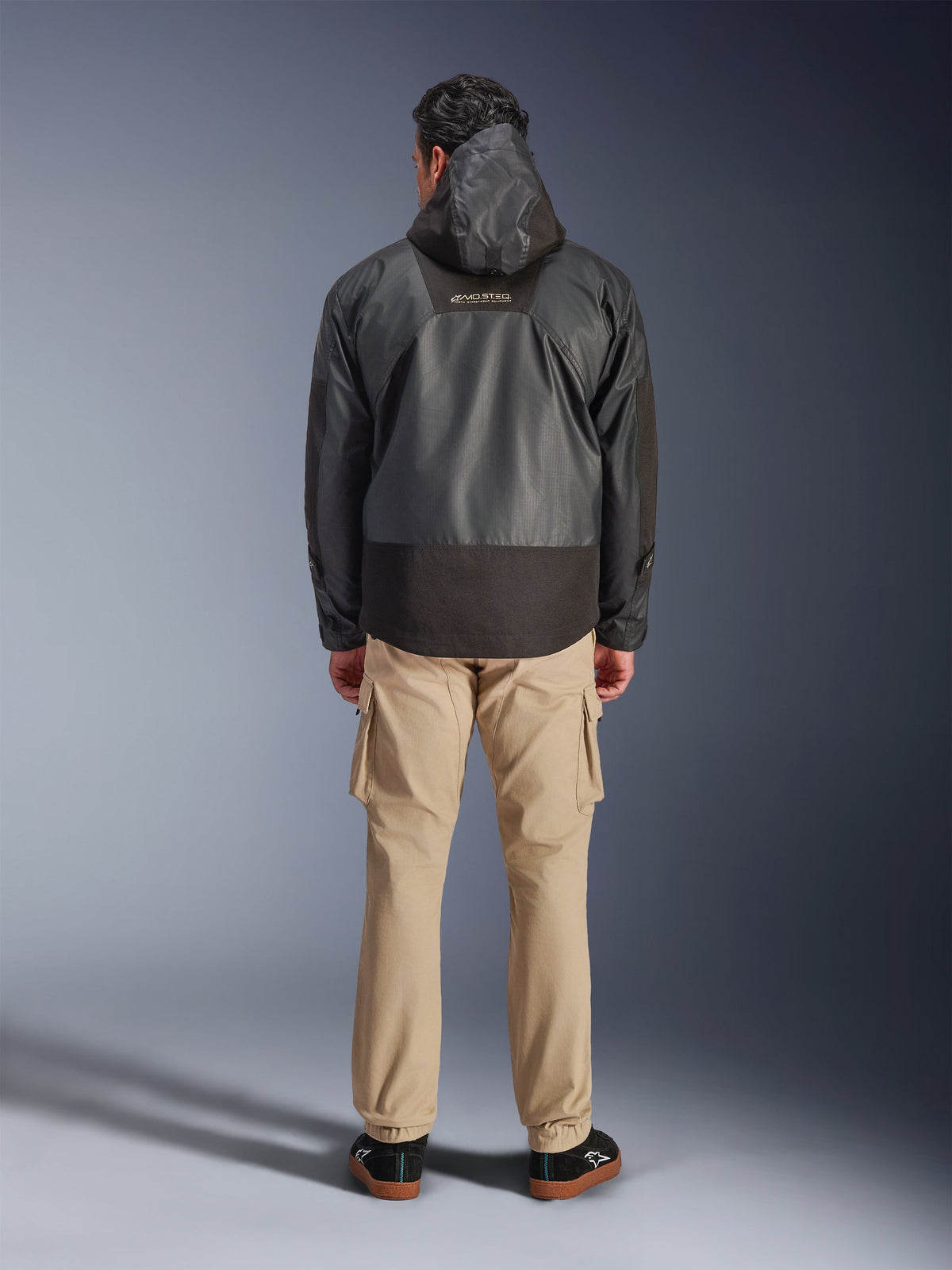 Veste imperméable Alpinestars Mospher WP, parka de moto noire vue de dos, arborant le logo MO.ST.EQ sur le haut du dos et une construction en tissu ripstop, portée avec un pantalon cargo beige et des baskets noires, style urbain.