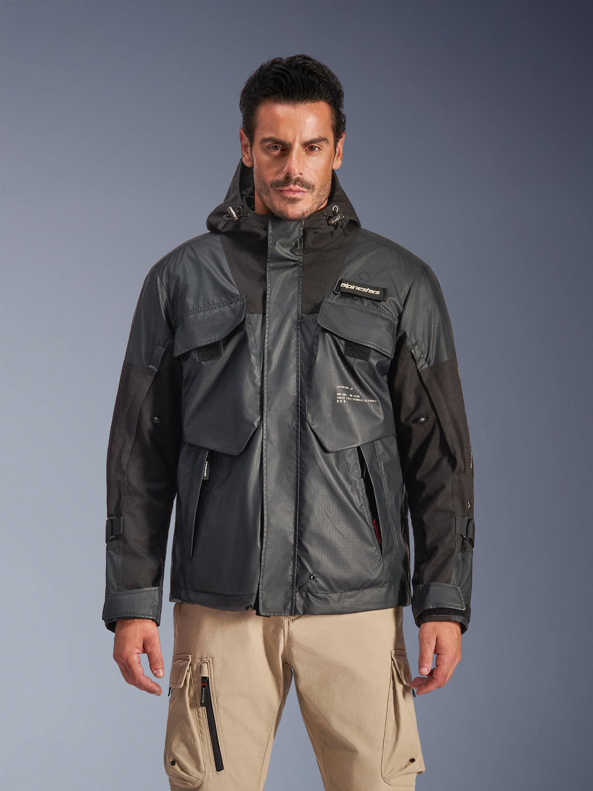 Alpinestars Mospher Waterproof Jacket, parka de moto urbaine pour homme, tissu ripstop noir et gris foncé, design à capuche avec plusieurs grandes poches utilitaires, portée par un mannequin avec un pantalon cargo kaki pour un look tactique