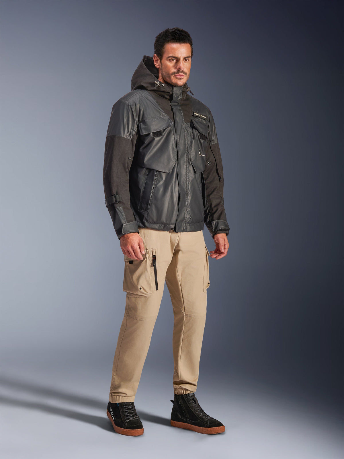 Modèle masculin portant la veste Alpinestars Mospher Waterproof noire, parka imperméable d'inspiration utilitaire avec design multi-poches et capuche, associée à un pantalon cargo beige et des baskets montantes noires, équipement de moto urbain.