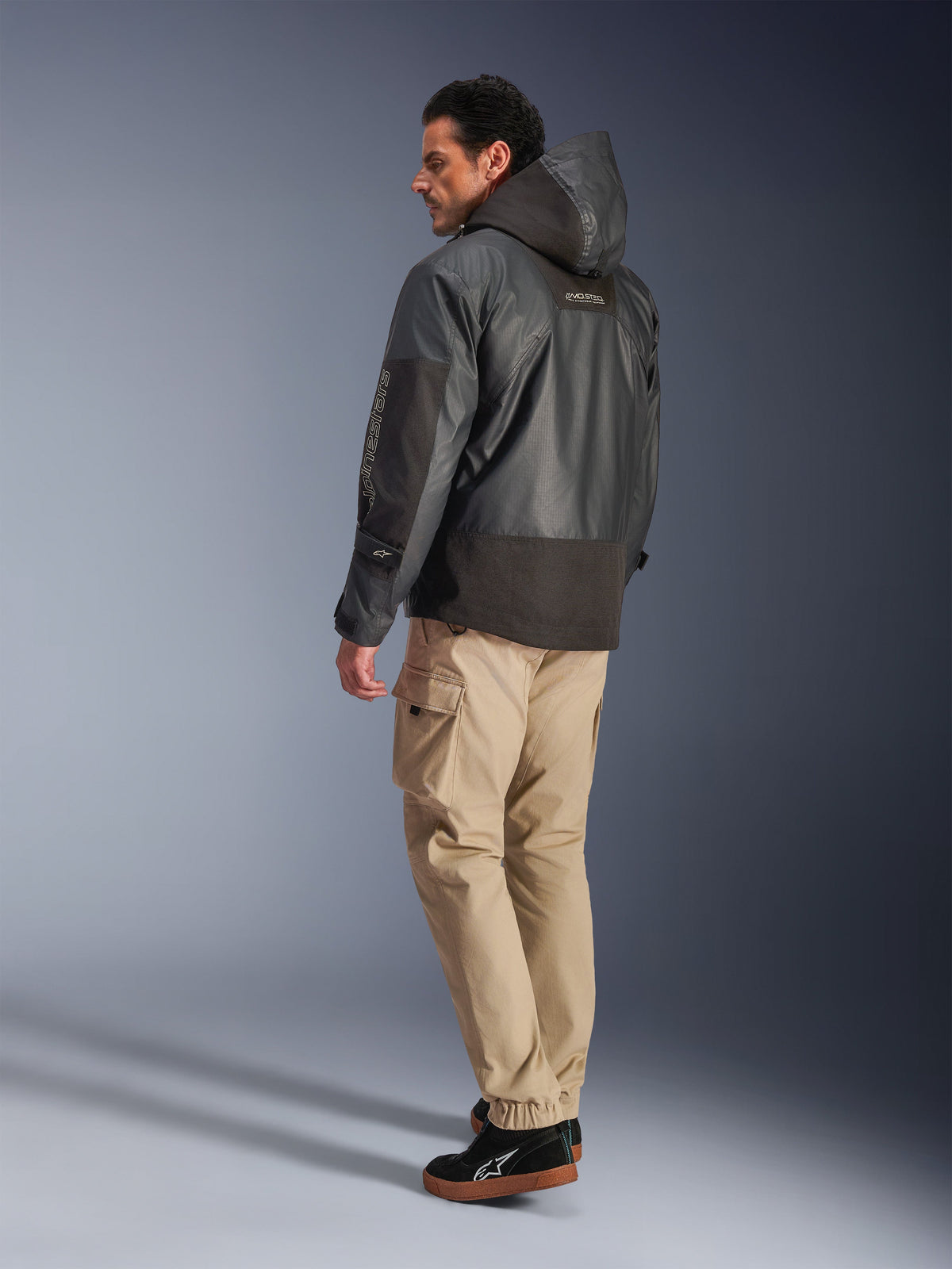 Homme portant la veste imperméable Alpinestars Mospher en noir, vue de profil arrière et latéral, avec pantalon cargo beige et baskets noires, design parka à capuche en tissu ripstop, marquage Alpinestars sur la manche, style moto urbain moderne
