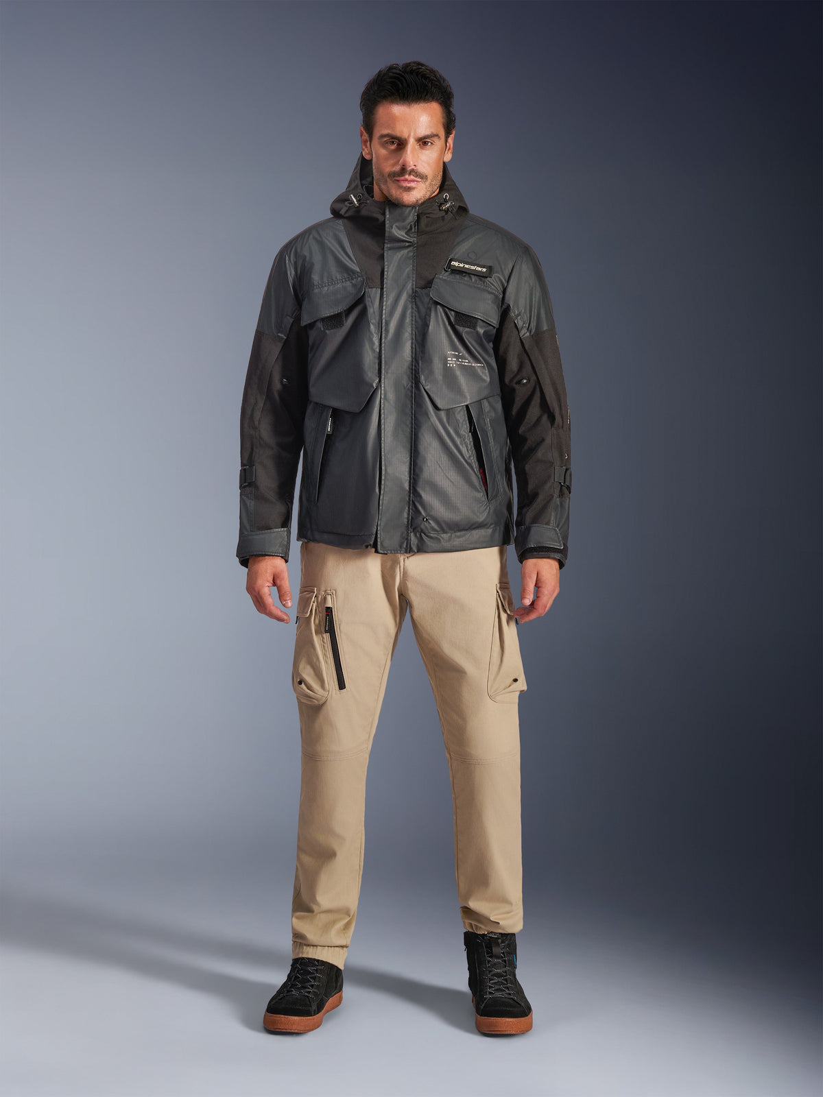 Modèle masculin portant la veste imperméable Alpinestars Mospher en noir, équipement de moto urbain contemporain, design utilitaire tactique avec de multiples poches fonctionnelles et une capuche, associé à un pantalon cargo beige et des chaussures noires, portrait de studio en pied.