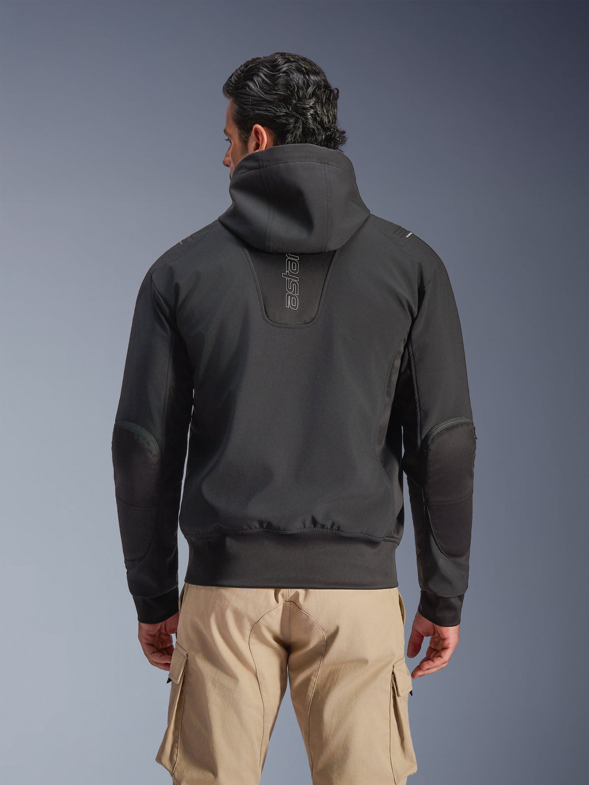 Veste imperméable Alpinestars Moblast, noire, vue arrière d'une veste de moto urbaine à capuche, avec logo « astars » vertical au dos, taille et poignets élastiqués, coudes renforcés et design technique moderne, portée avec un pantalon cargo beige
