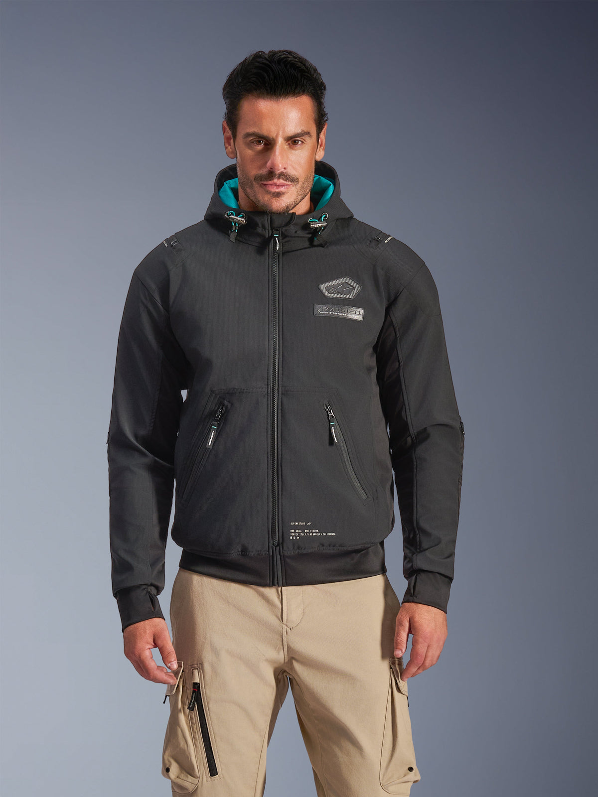 Alpinestars Moblast Waterproof Jacket, veste de moto urbaine pour homme, noire avec doublure de capuche turquoise, poches zippées 3D et design à capuche, portée avec un pantalon cargo beige, tenue de moto urbaine moderne