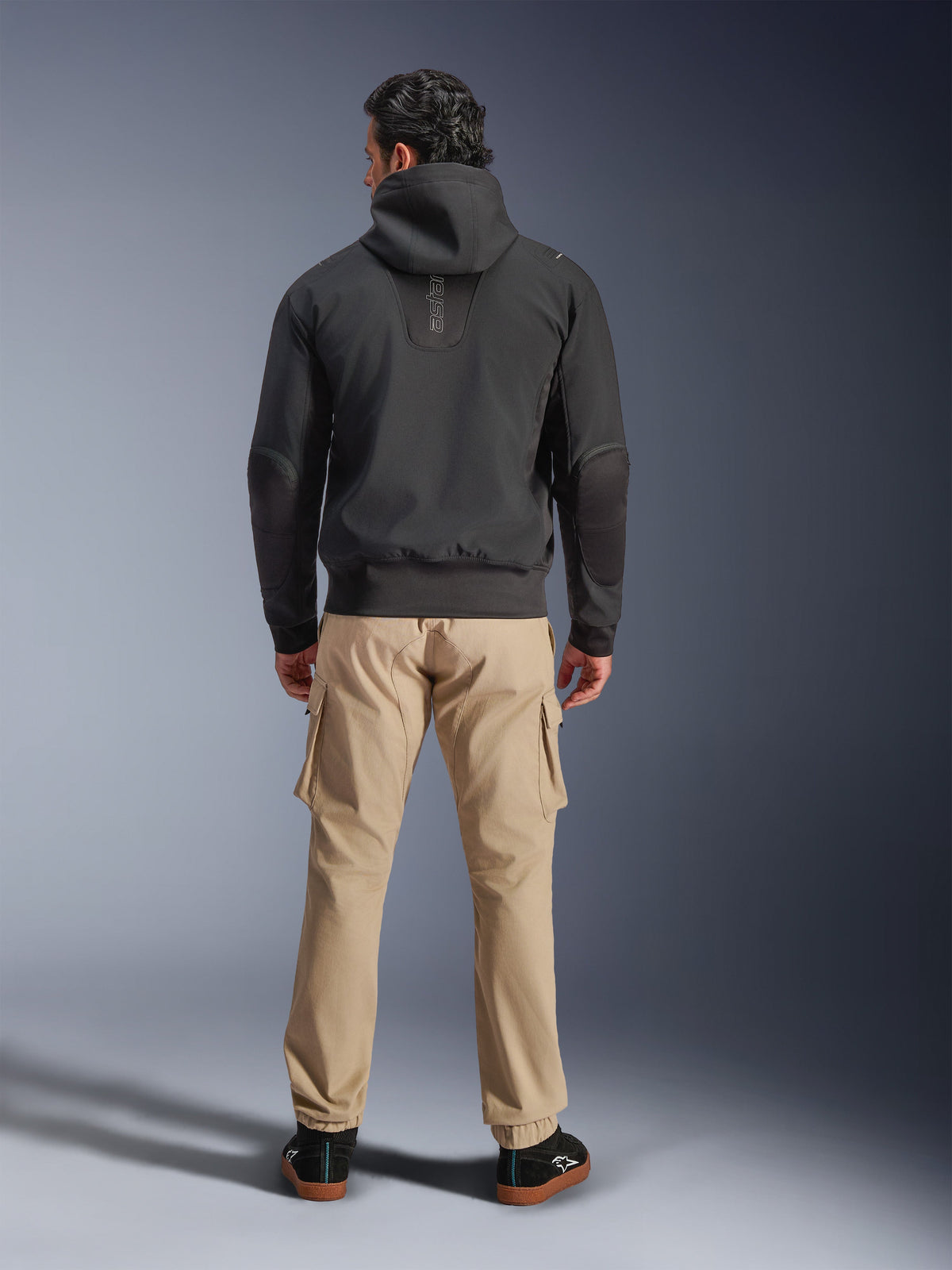 Veste imperméable Alpinestars Moblast, noir, vue arrière d'un hoodie de moto urbain, porté avec un pantalon cargo beige et des baskets noires, design minimaliste avec marquage vertical « astars » sur le haut du dos, équipement de conduite contemporain toutes saisons