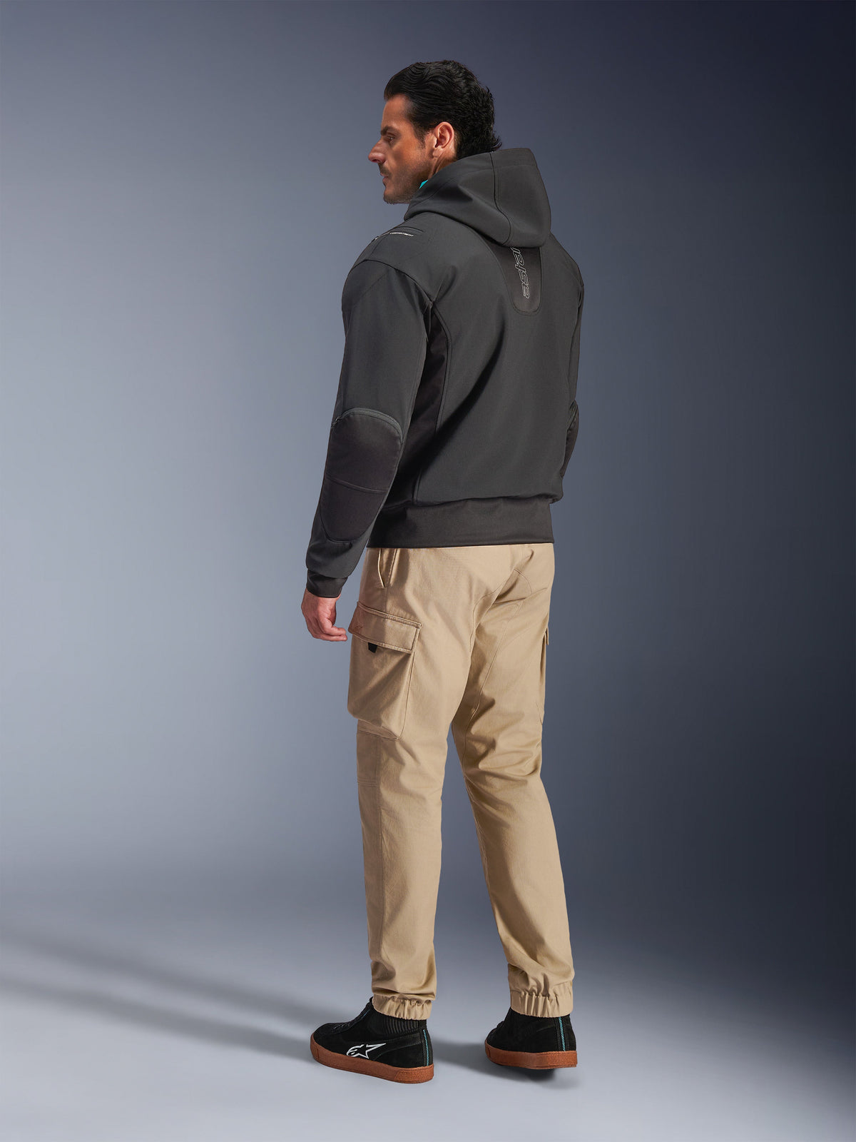 Veste imperméable Alpinestars Moblast, hoodie technique de moto noir, vue de côté et de dos, porté avec un pantalon cargo beige et des baskets noires, design à capuche, construction en textile technique et esthétique urbaine moderne