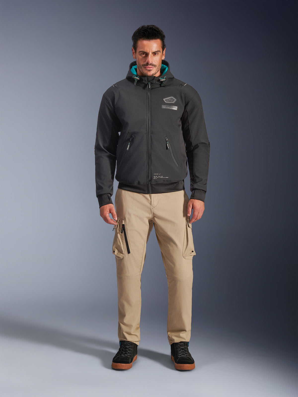 Veste imperméable Alpinestars Moblast, hoodie de moto urbain noir avec doublure de capuche bleu sarcelle, porté avec un pantalon cargo beige et des bottes montantes noires, vêtement de moto contemporain toutes saisons