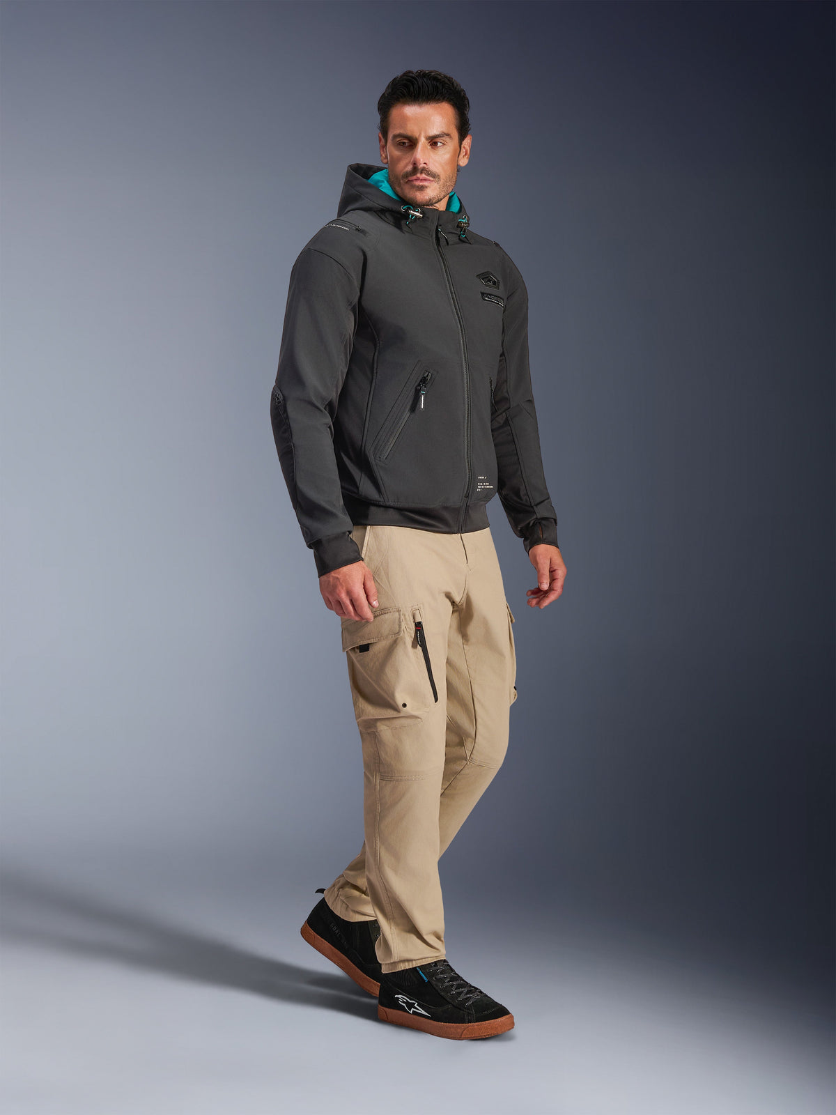 Mannequin homme portant la veste imperméable Alpinestars Moblast en noir, hoodie de moto urbain avec capuche doublée de bleu sarcelle, associé à un pantalon cargo kaki et des baskets noires, design minimaliste moderne avec tissu technique extensible pour les trajets par tous les temps