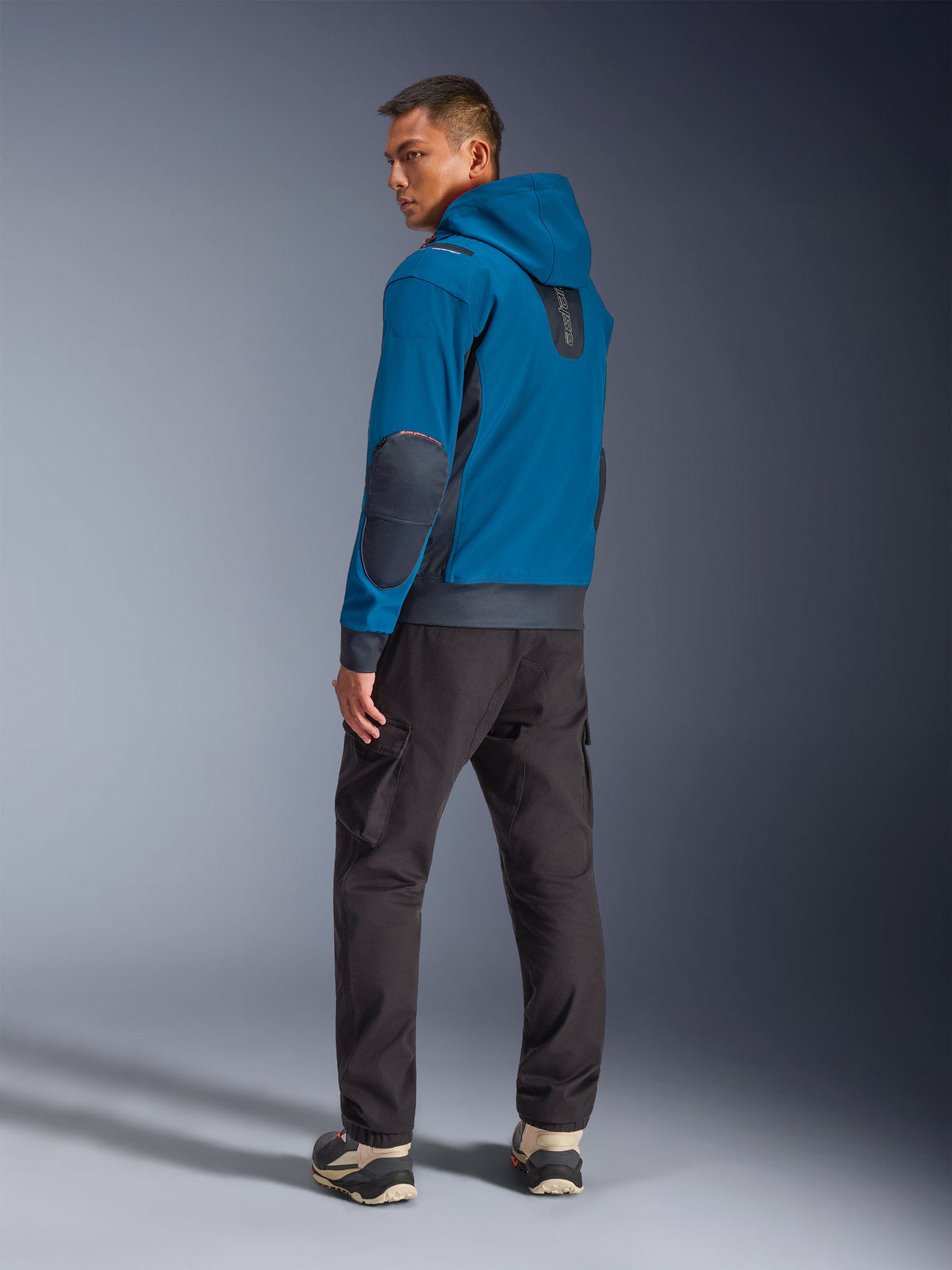 Veste imperméable Alpinestars Moblast en Dark Petrol et Dark Navy, vue de dos sur mannequin montrant la conception à capuche, les renforts aux coudes et les inserts latéraux bleu marine foncé, associée à un pantalon cargo sombre et des baskets techniques, tenue de moto urbaine moderne.