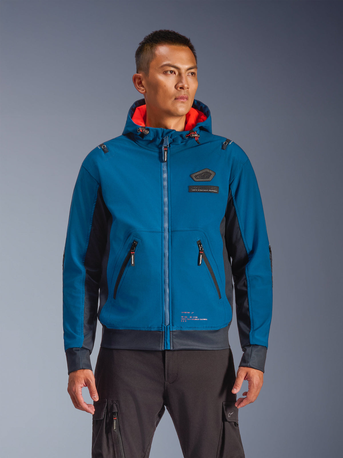 Veste imperméable Alpinestars Moblast portée par un mannequin, bleu et bleu marine foncé, veste de moto urbaine, design à capuche avec doublure intérieure rouge, fermetures éclair noires et détail de patch sur la poitrine, équipement de moto d'inspiration streetwear