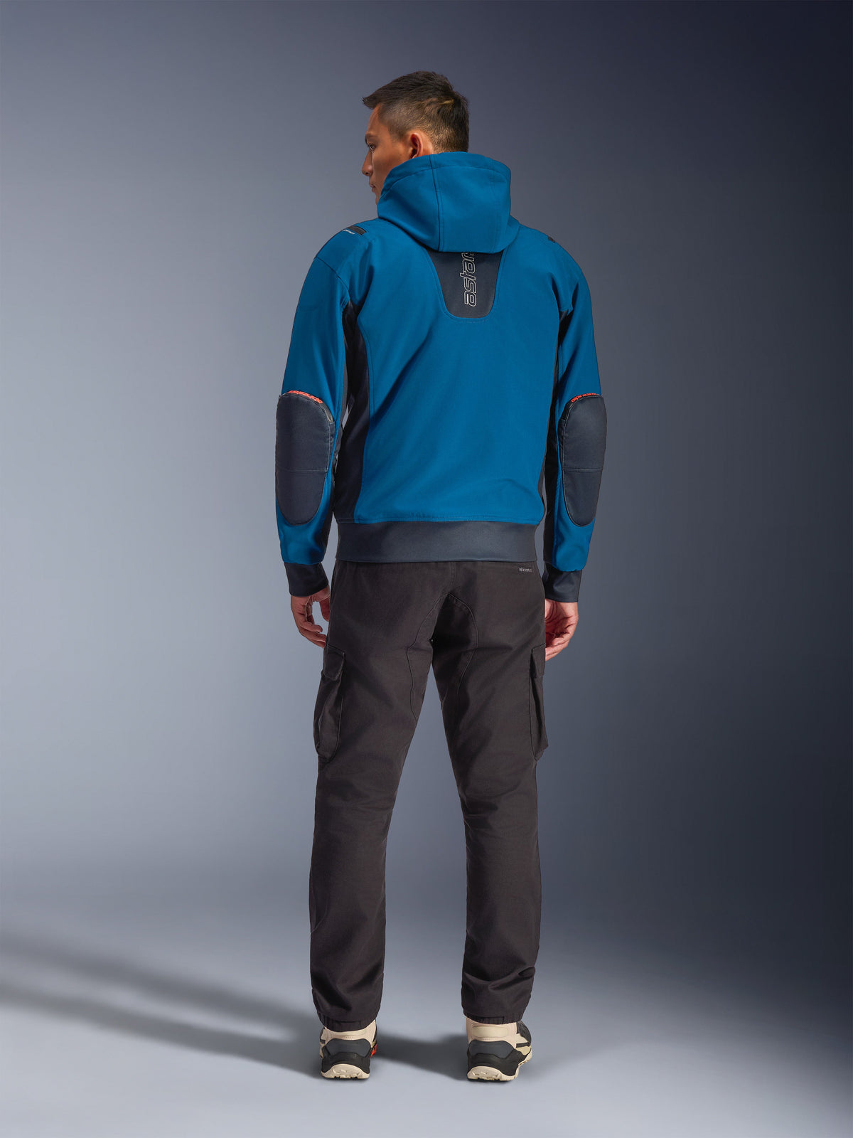 Veste imperméable Alpinestars Moblast, veste de moto urbaine, bleu pétrole et bleu marine foncé, vue arrière montrant la capuche et les renforts aux coudes, portée avec un pantalon cargo sombre, équipement de conduite technique toutes saisons