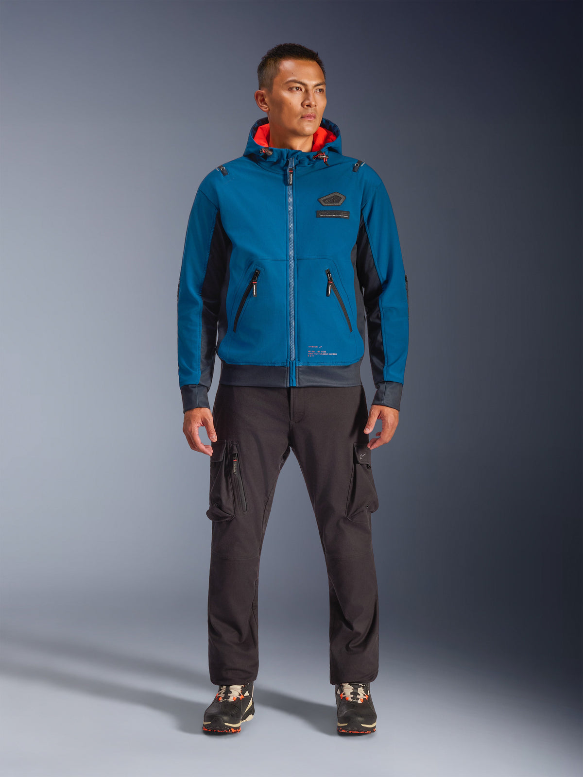 Veste imperméable Alpinestars Moblast, veste de moto à capuche bleu pétrole foncé et bleu marine foncé avec doublure intérieure orange, portée par un mannequin homme avec un pantalon cargo foncé et des baskets noires, équipement de conduite urbain moderne