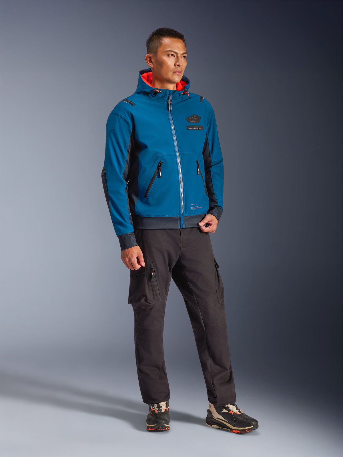 Mannequin homme portant la veste imperméable Alpinestars Moblast en Dark Petrol et Dark Navy, dotée d'une capuche avec intérieur orange vif, associée à un pantalon cargo noir et des baskets, équipement de moto urbain moderne.