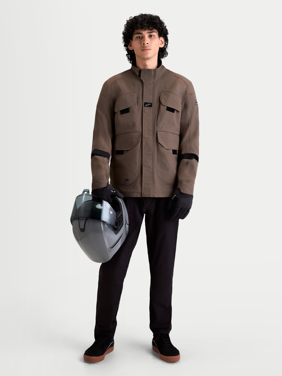 Alpinestars Altan Drystar Veste, veste de moto urbaine et contemporaine pour homme en marron marron, vue de face complète sur un modèle portant un pantalon noir, des gants et tenant un casque, construction textile imperméable avec poches cargo