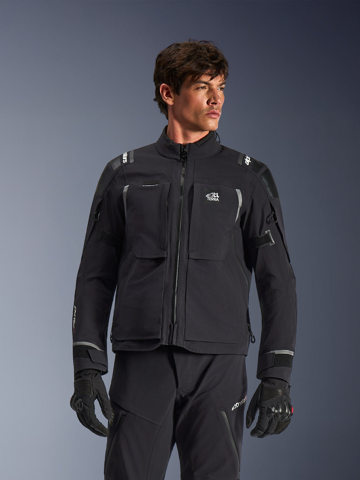 Alpinestars Andes Pro Drystar® XF Jacket, black and dark gray motorcycle adventure touring jacket, worn with matching pants and gloves, featuring technical laminated shell construction, ALL TERRA chest logo, reflective details, and adjustable cuffs