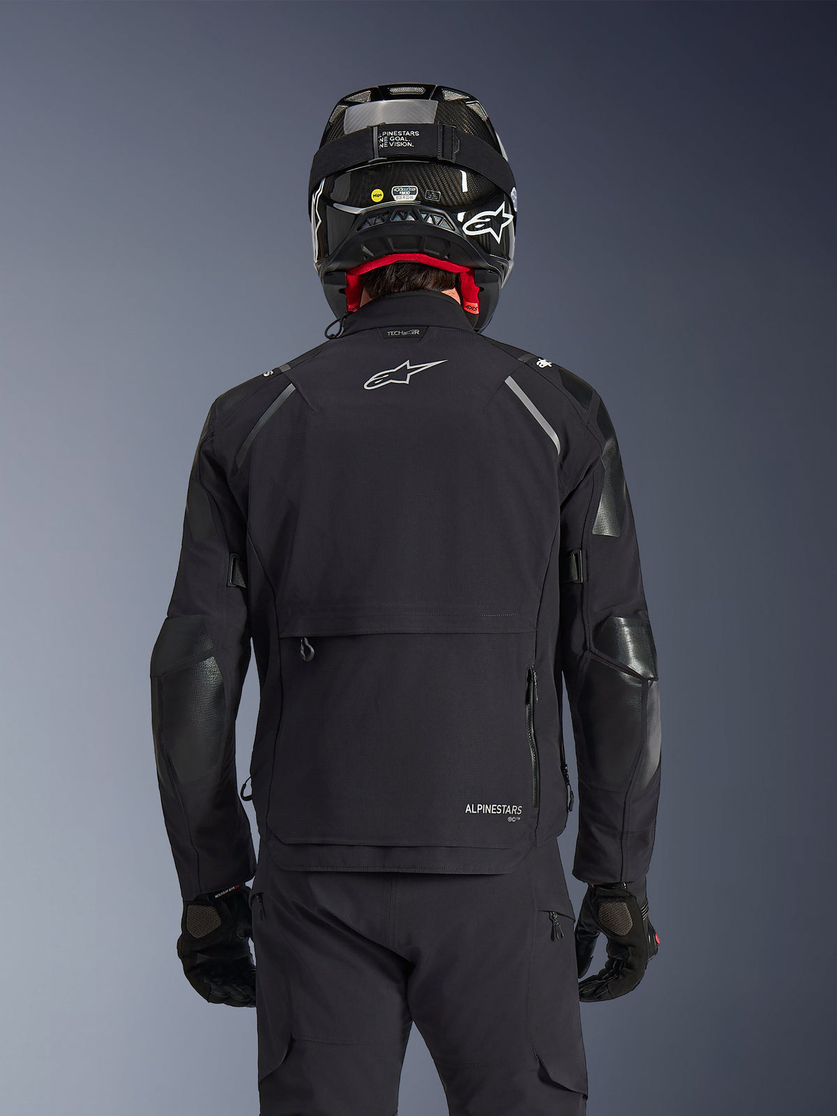 Alpinestars Andes Pro Drystar® XF Jacket in Black, rear view on a rider wearing a carbon fiber helmet, featuring a large rear storage compartment and white Alpinestars logo, technical all-season adventure motorcycle gear.
