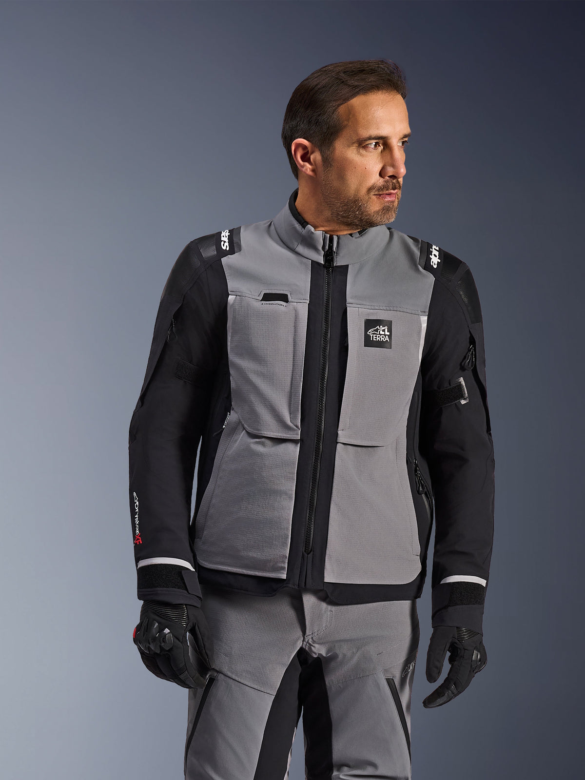 Veste Alpinestars Andes Pro Drystar® XF en gris foncé et noir, équipement de moto d'aventure, portée par un homme avec pantalon gris et gants noirs assortis, dotée d'une coque laminée 3 couches, de poches utilitaires et du logo « Terra » sur la poitrine