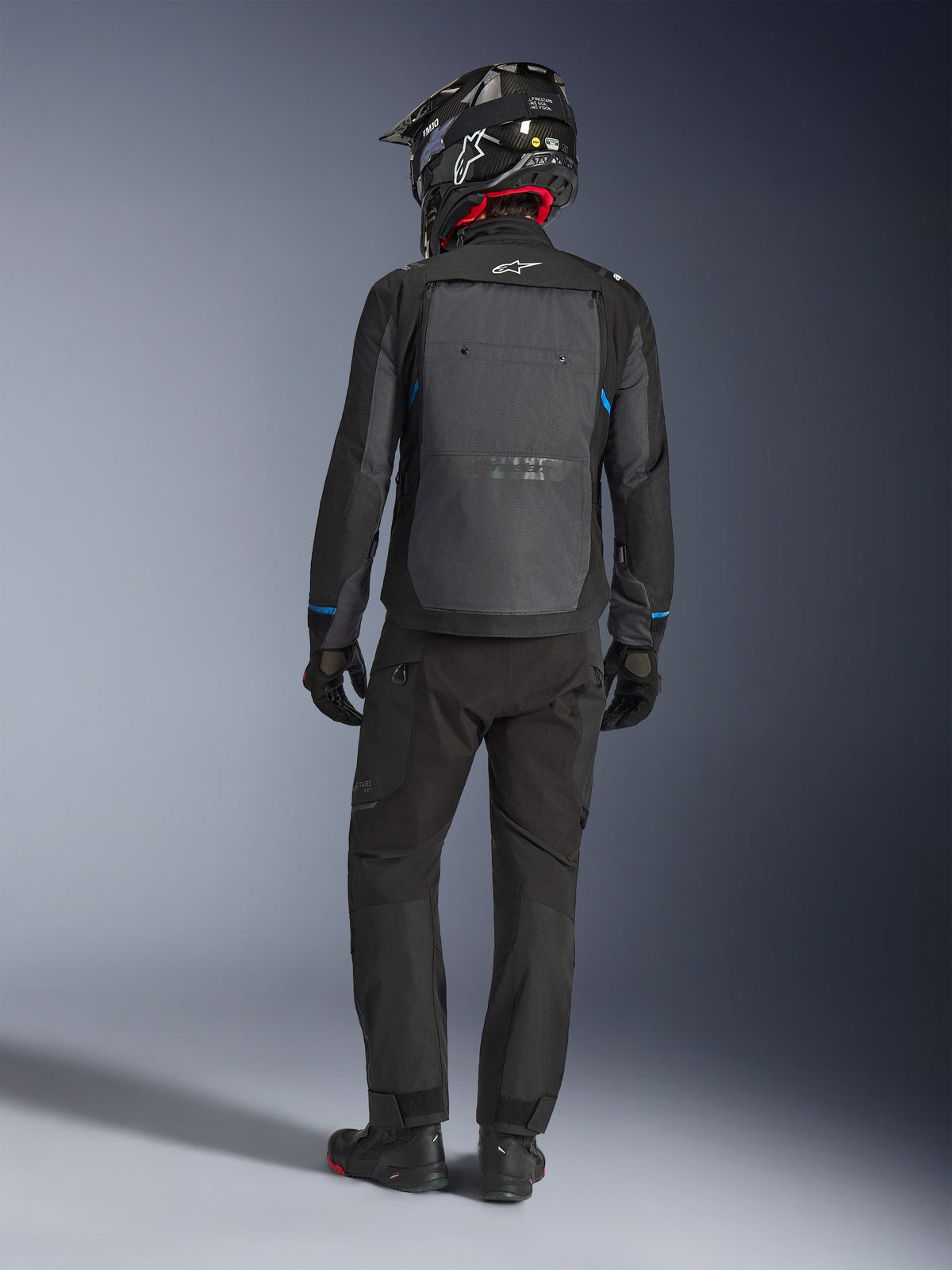 Veste Alpinestars Cusco Drystar®, noir et gris foncé, vue arrière d'un motard en tenue complète d'aventure touring, grand panneau arrière gris avec logo Alpinestars, accents bleus sur les manches, portée avec un pantalon noir et un casque assortis.