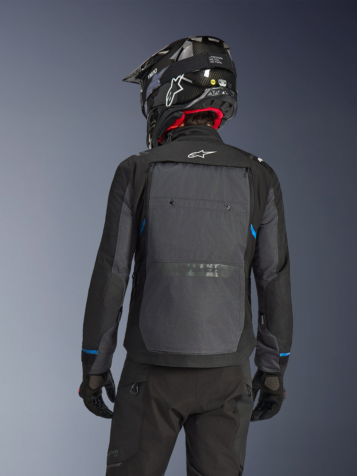 Pilote vu de dos portant la veste Alpinestars Cusco Drystar® en Noir et Gris Foncé, dotée d'un grand panneau de ventilation arrière gris et d'un logo blanc, accompagnée d'un casque tout-terrain noir et d'un équipement de moto noir pour l'adventure touring.