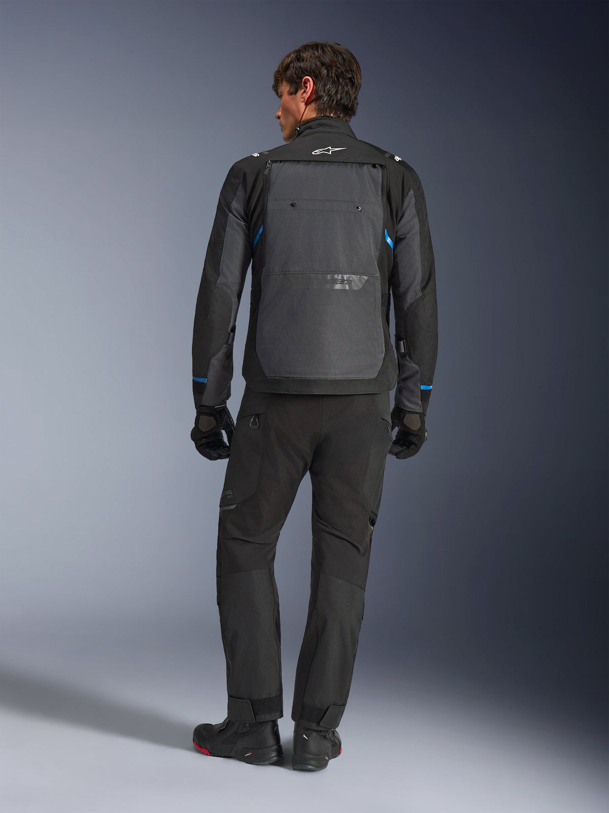 Veste Alpinestars Cusco Drystar®, noir et gris foncé avec des accents bleus, vue de dos, portée avec un pantalon d'aventure et des gants noirs assortis, dotée d'un large panneau dorsal gris et d'un logo blanc sur le col, conçue pour la route et le touring d'aventure