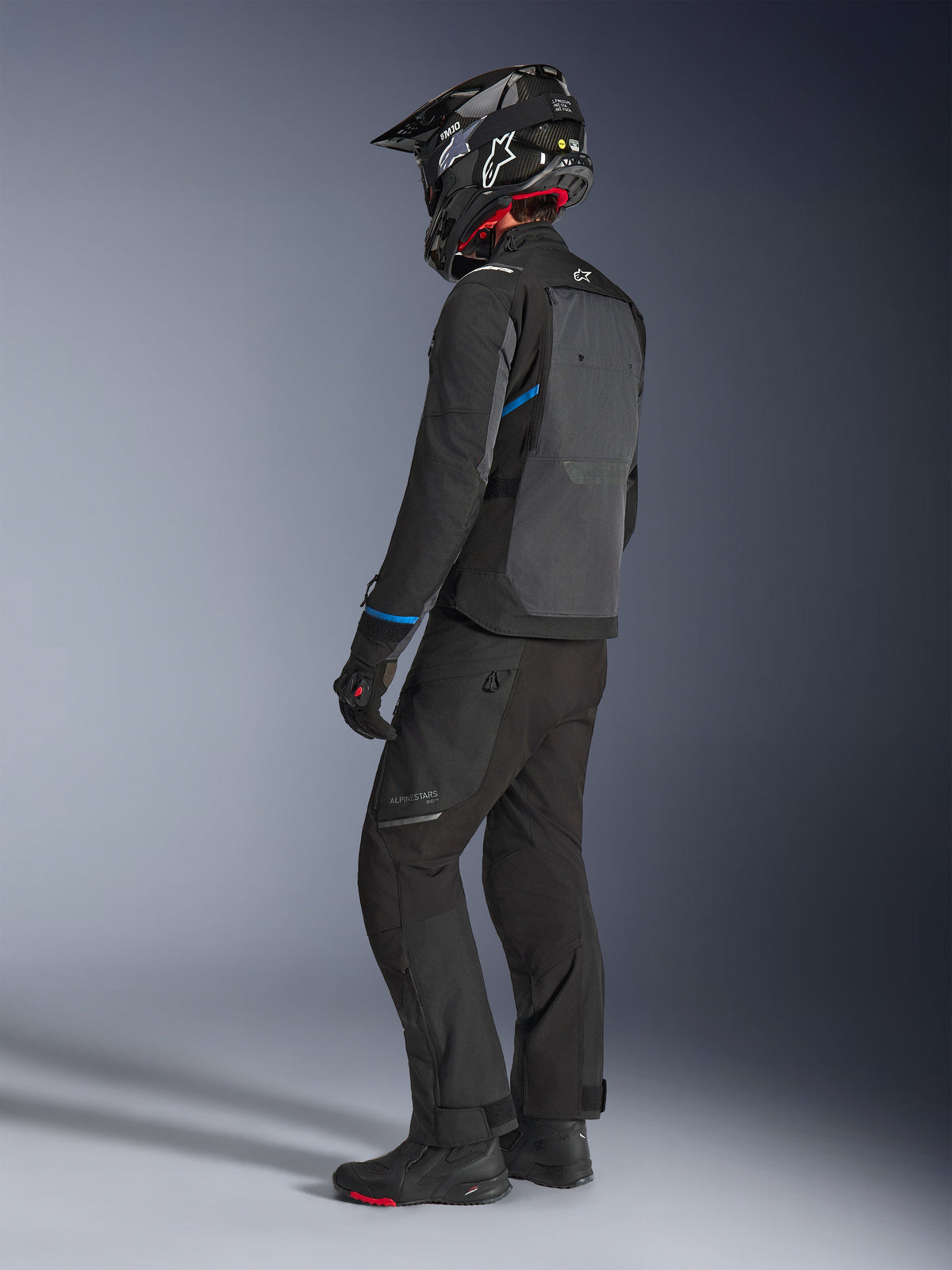 Motard portant la veste Alpinestars Cusco Drystar® en noir et gris foncé avec accents bleus, associée à un pantalon de moto noir, un casque SM10 en fibre de carbone et des gants noirs, vue de dos montrant le large panneau de ventilation et le design adventure touring.