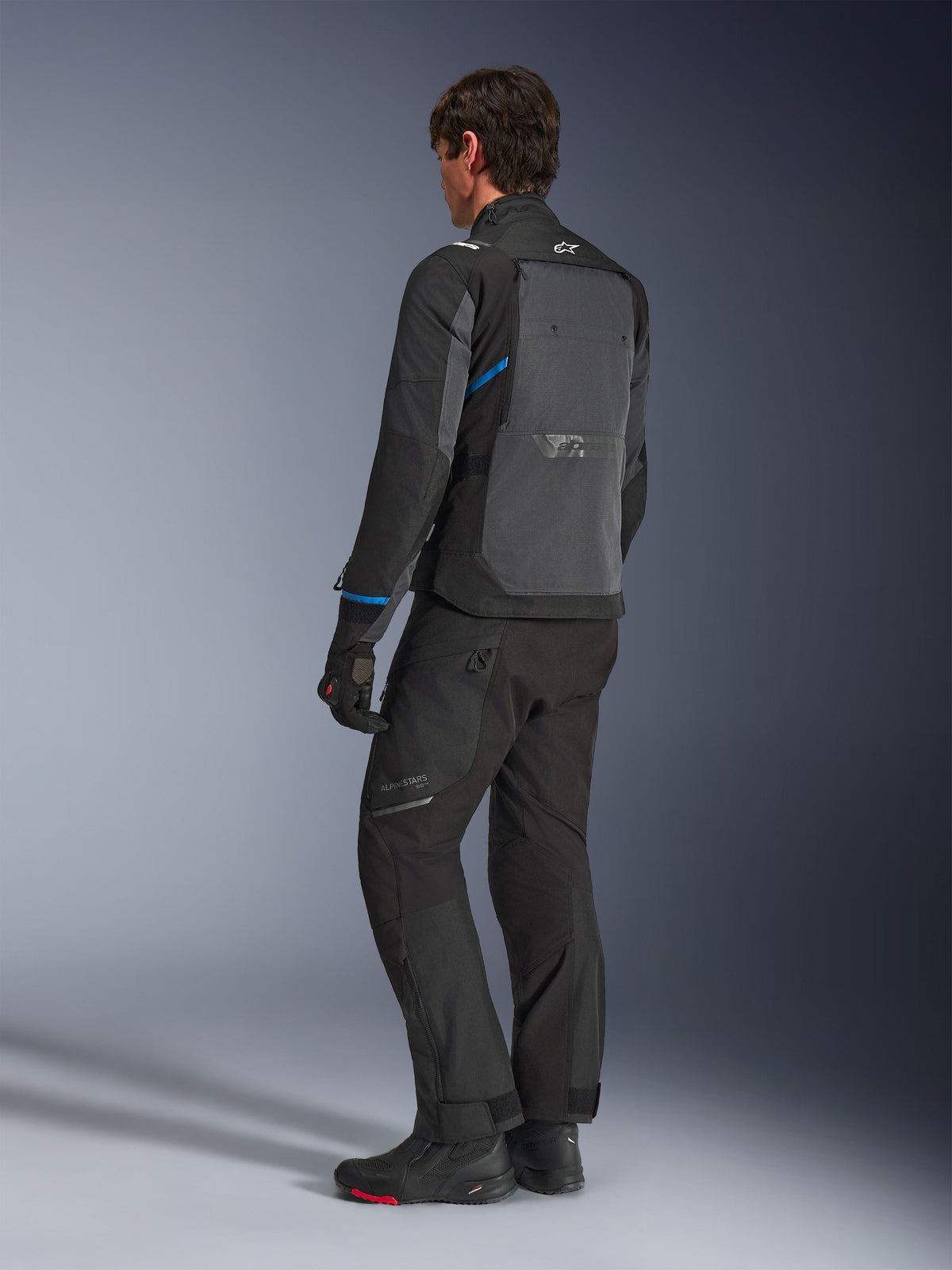 Veste Alpinestars Cusco Drystar®, noir et gris foncé, vue arrière de trois-quarts montrant le grand panneau de ventilation arrière et les accents bleus sur les manches, portée avec un pantalon et des bottes de moto noirs pour une tenue de touring complète.