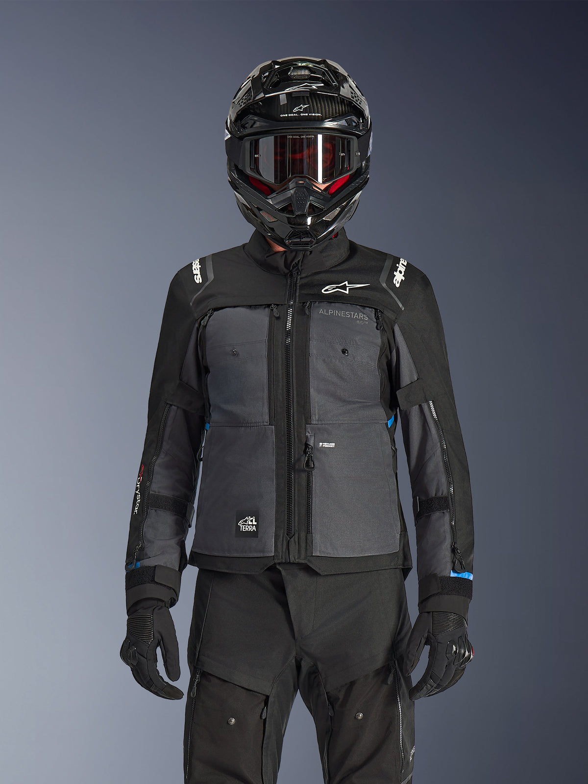 Veste Alpinestars Cusco Drystar®, noir et gris foncé, veste de moto adventure touring portée avec un pantalon technique noir assorti et un casque dual-sport, dotée d'un design bicolore avec empiècements en ripstop et marquage Alpinestars