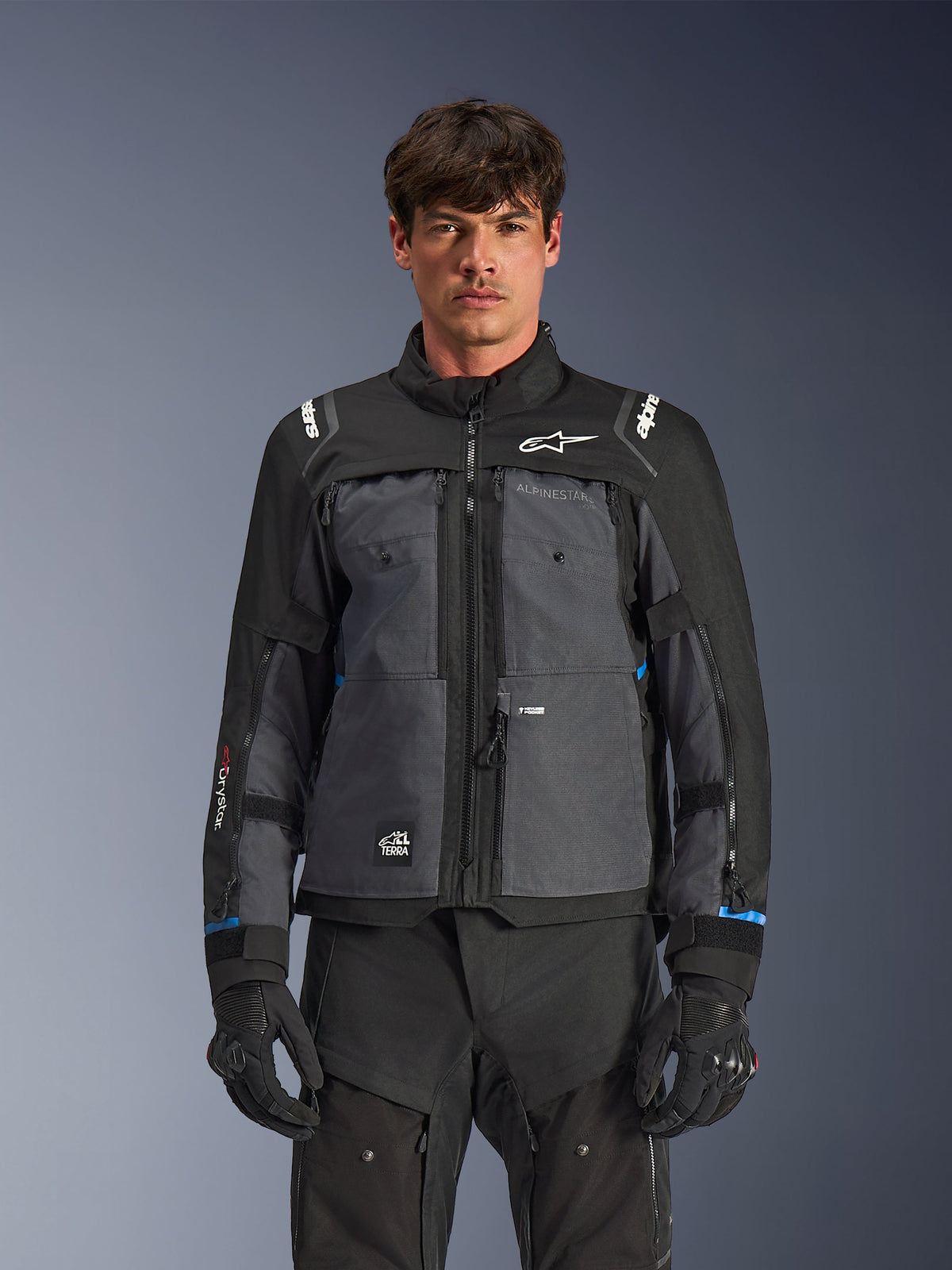 Veste Alpinestars Cusco Drystar®, veste de moto touring, noir et gris foncé, vue de face sur modèle, avec logos Alpinestars sur la poitrine et les épaules, marquage Drystar® sur la manche, design technique multi-poches avec zips de ventilation, associée à un équipement de moto noir