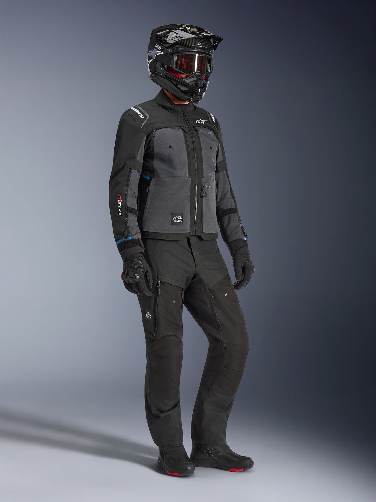 Mannequin portant la veste Alpinestars Cusco Drystar® en Noir et Gris foncé, associée à une tenue de moto complète incluant un casque noir, un pantalon technique assorti, des gants et des bottes, avec une construction en ripstop et softshell pour la route et l'adventure touring