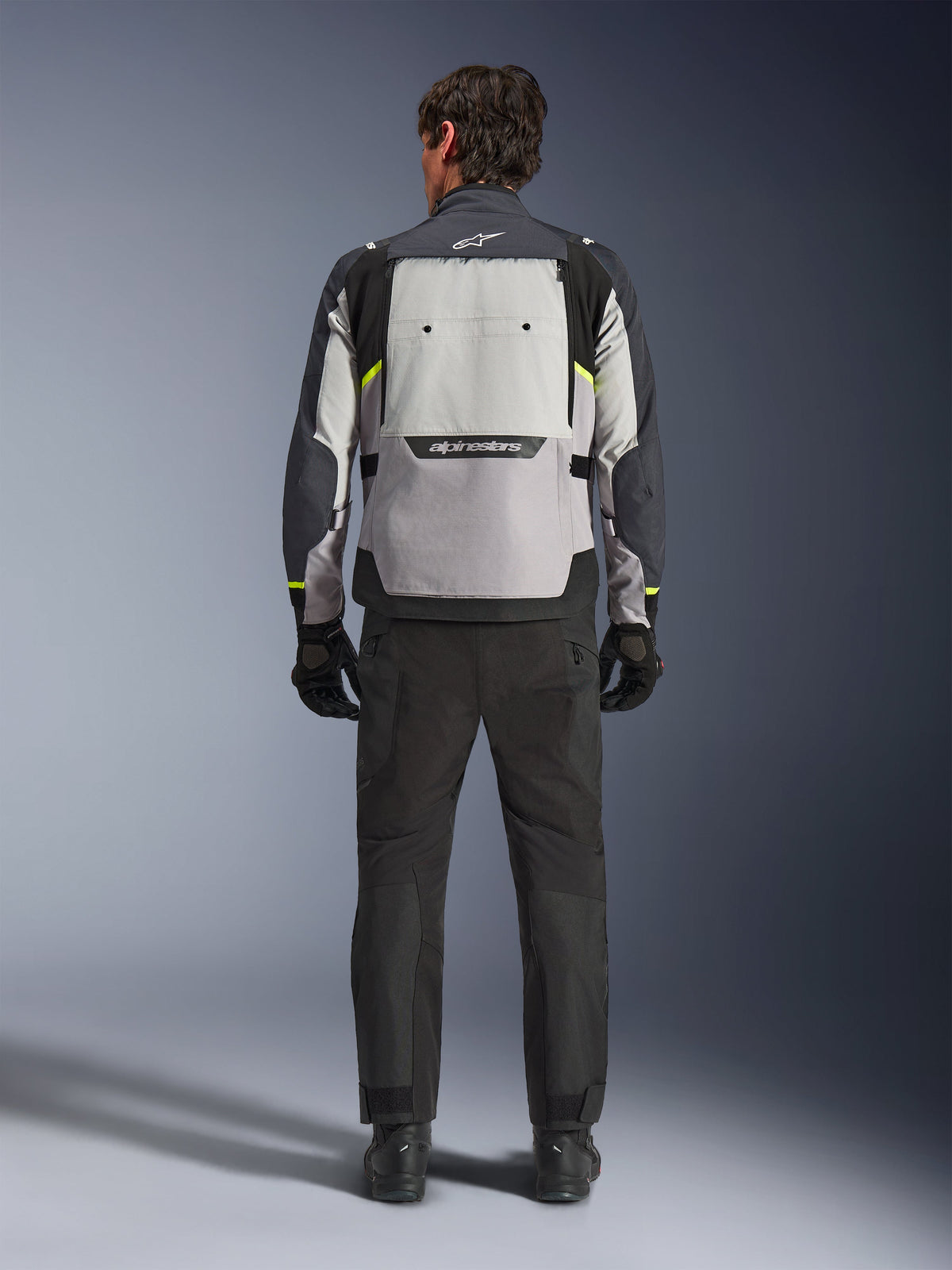 Vue arrière d'un motard portant la veste Alpinestars Cusco Drystar® en Ice Gray, Mid Gray et Deep Gray, associée à un pantalon et des gants de moto noirs, mettant en évidence le large panneau de ventilation arrière et le logo Alpinestars.