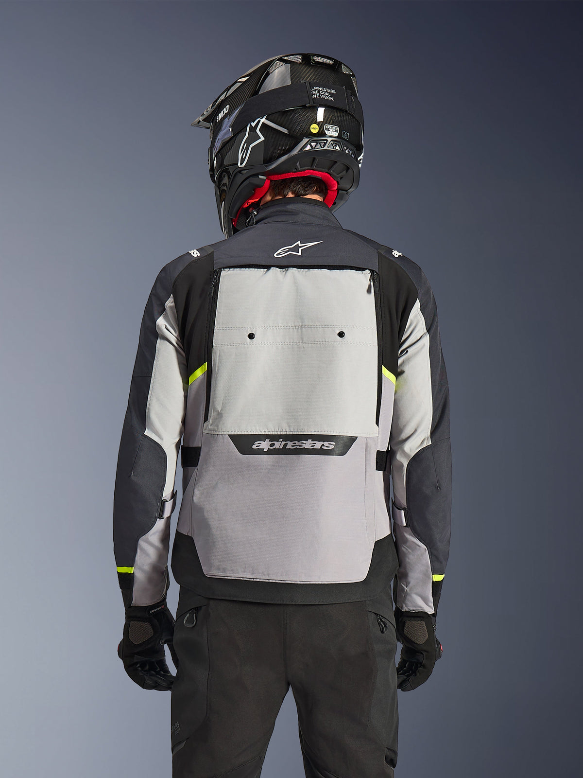 Veste Alpinestars Cusco Drystar®, vue arrière, coloris Ice Gray, Mid Gray et Deep Gray, dotée d'un large panneau de ventilation dorsal et d'accents jaune haute visibilité, portée avec un casque d'aventure noir et un pantalon noir.