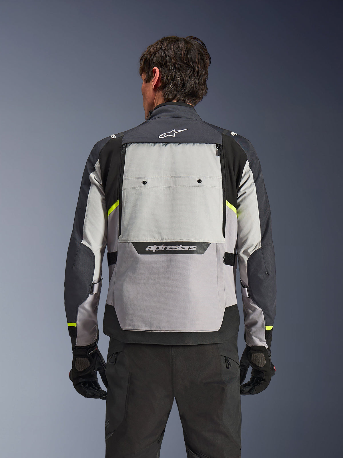 Vue arrière d'un motard portant la veste Alpinestars Cusco Drystar® en Gris Glace, Gris Moyen et Gris Foncé, présentant des accents jaune néon, un large panneau de ventilation dorsal et un marquage Alpinestars audacieux, associée à un pantalon et des gants de moto noirs.